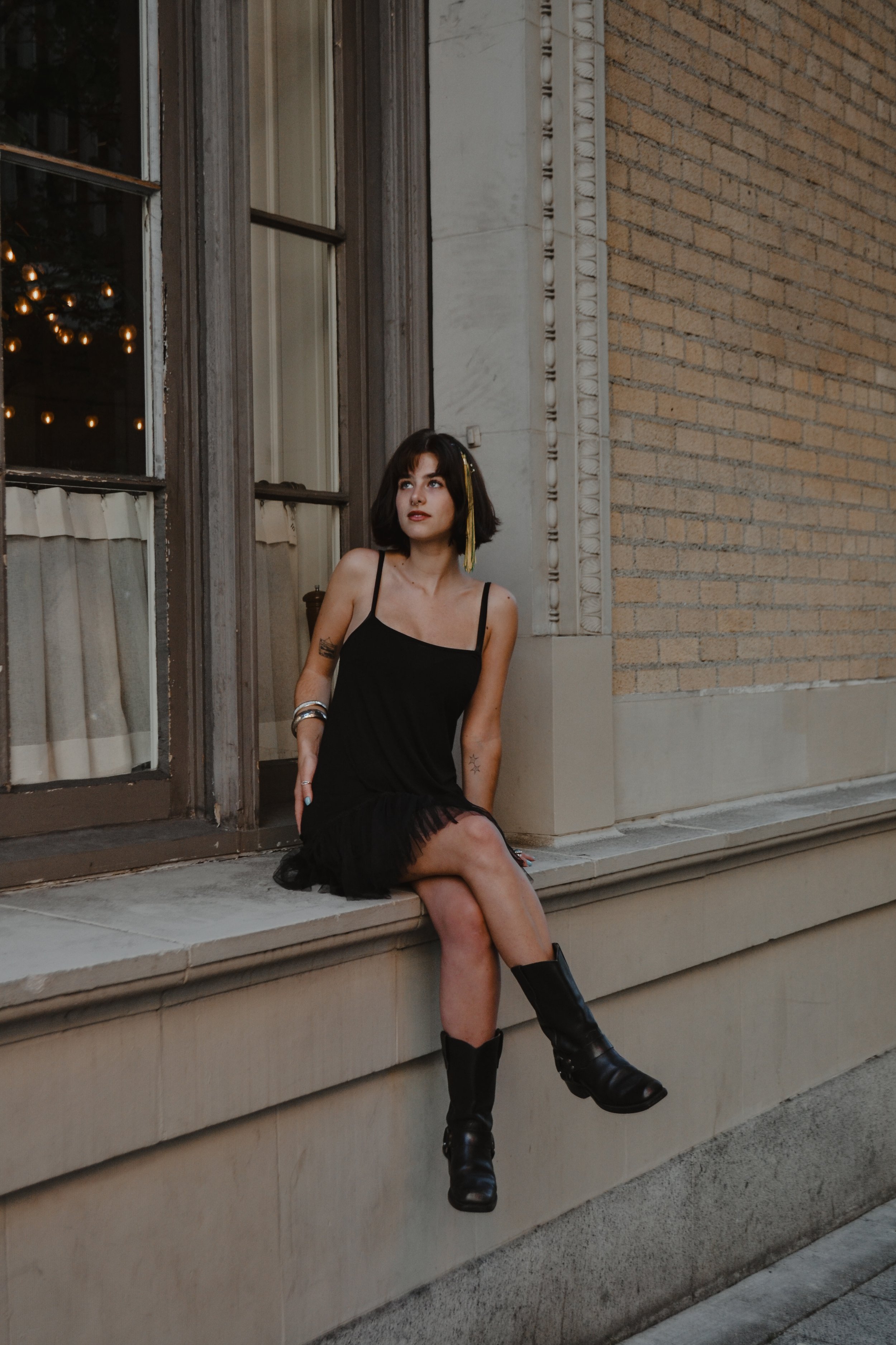 A young woman with dark hair and tattoos is sitting on a concrete ledge outside a building with brick and stone exterior. She is wearing a black dress, black boots, and has a yellow ribbon in her hair. The woman is looking upwards with a neutral expr
