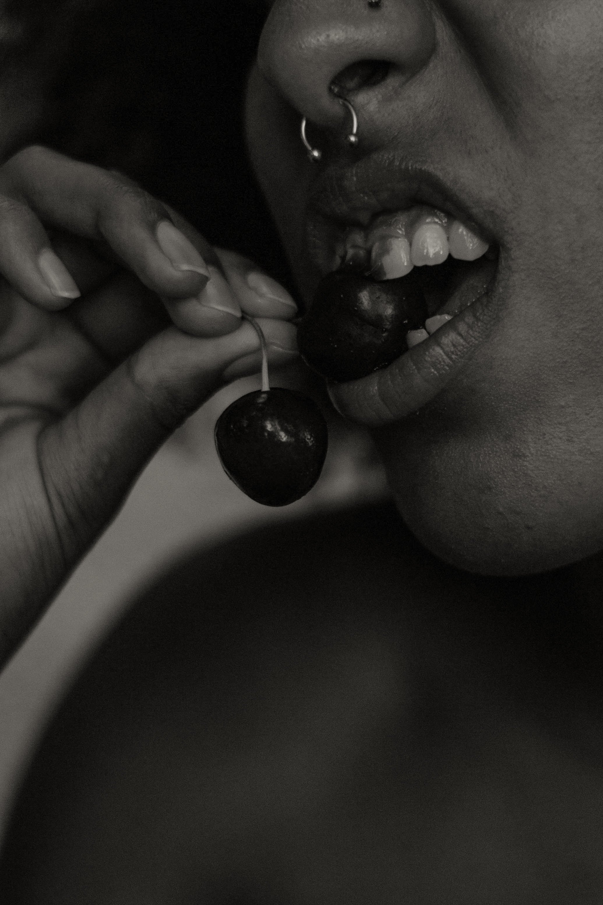 Close-up black and white photo of person with nose piercing, showing lips and teeth, holding a cherry near their mouth.