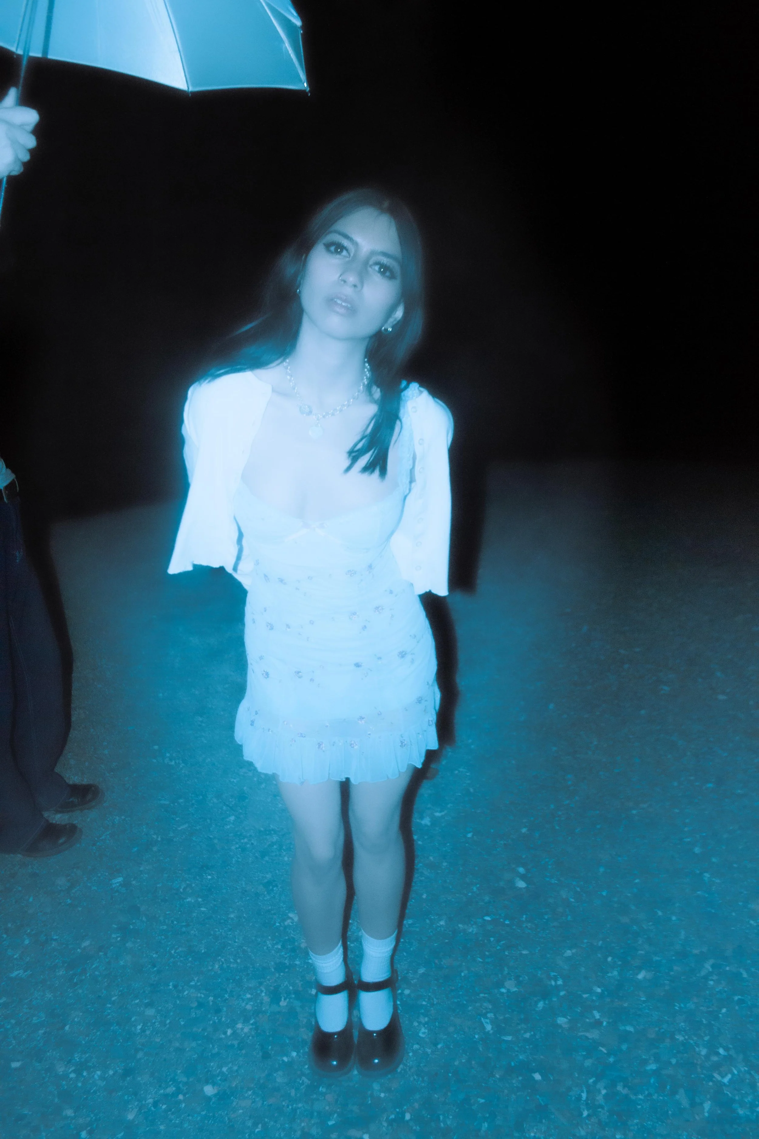 A young woman standing outdoors at night, holding a blue umbrella. She is wearing a white dress with floral patterns, white socks, and black shoes. She has long dark hair, earrings, and a necklace. The background is dark and she is illuminated by a n