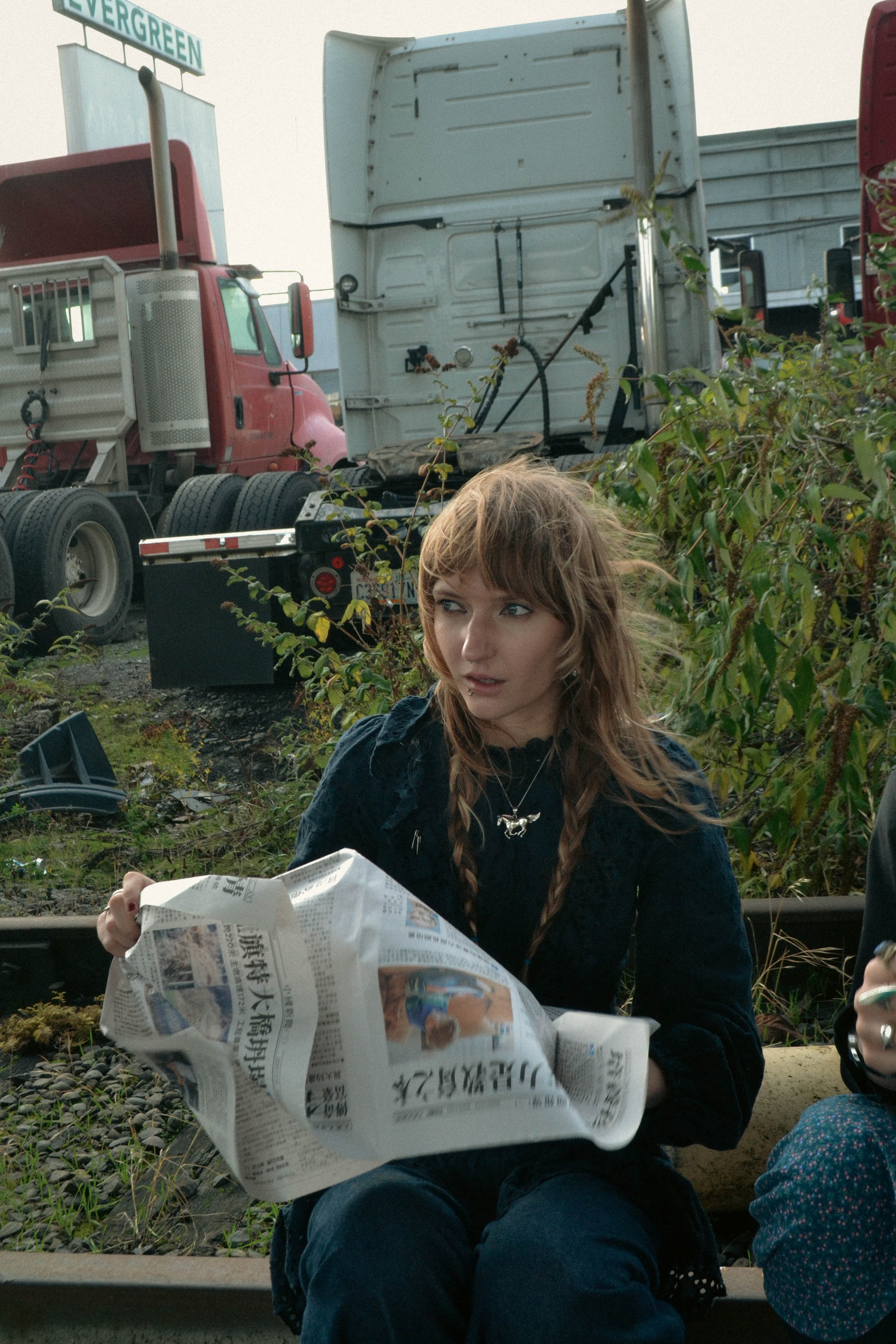 A young woman with brown hair and braided pigtails sitting on the ground, holding and reading a newspaper, with greenery and trucks in the background.