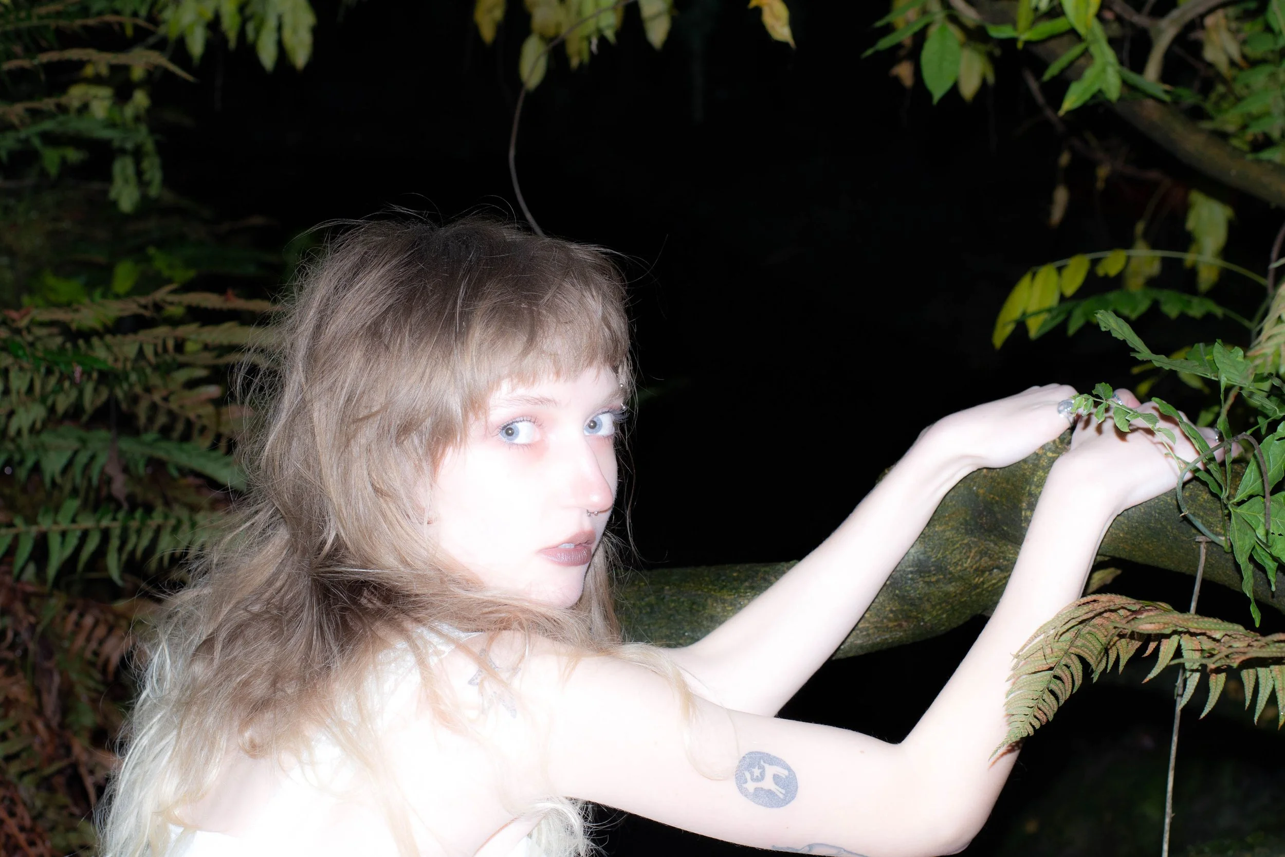 A person with long blonde hair and light skin reaching out in a green, leafy environment at night.