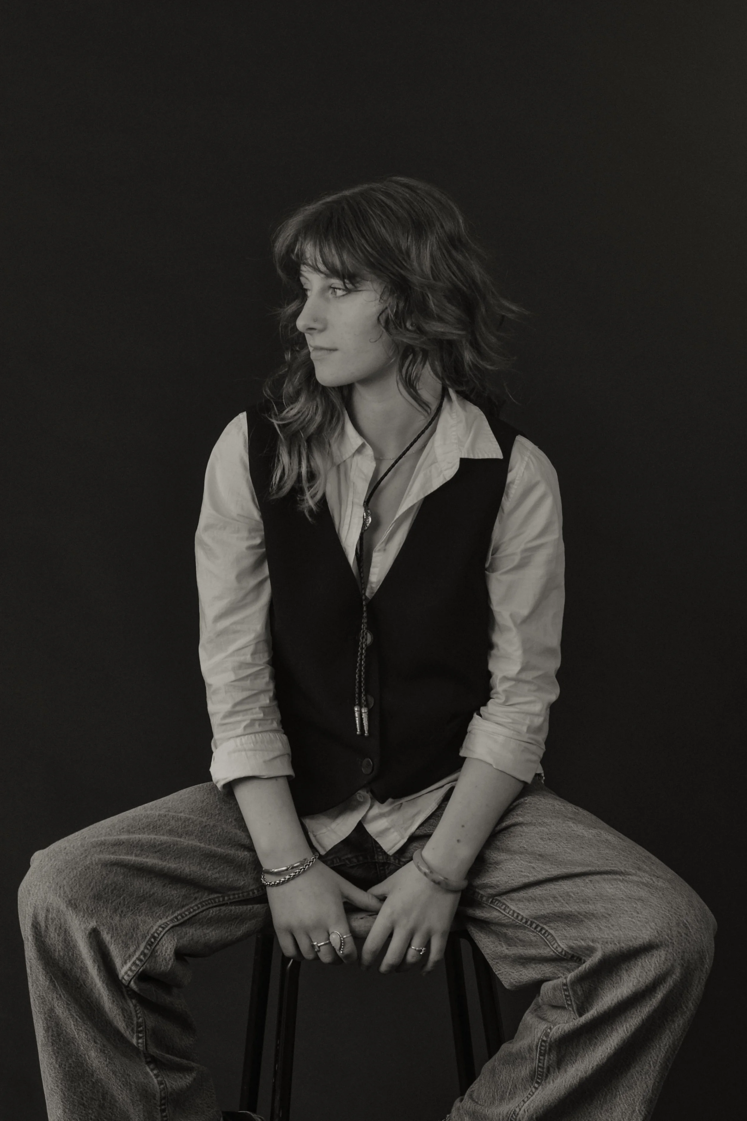 A woman sitting on a chair, wearing a collared shirt under a vest and baggy jeans, with wavy hair and jewelry, looking to her left against a plain dark background.