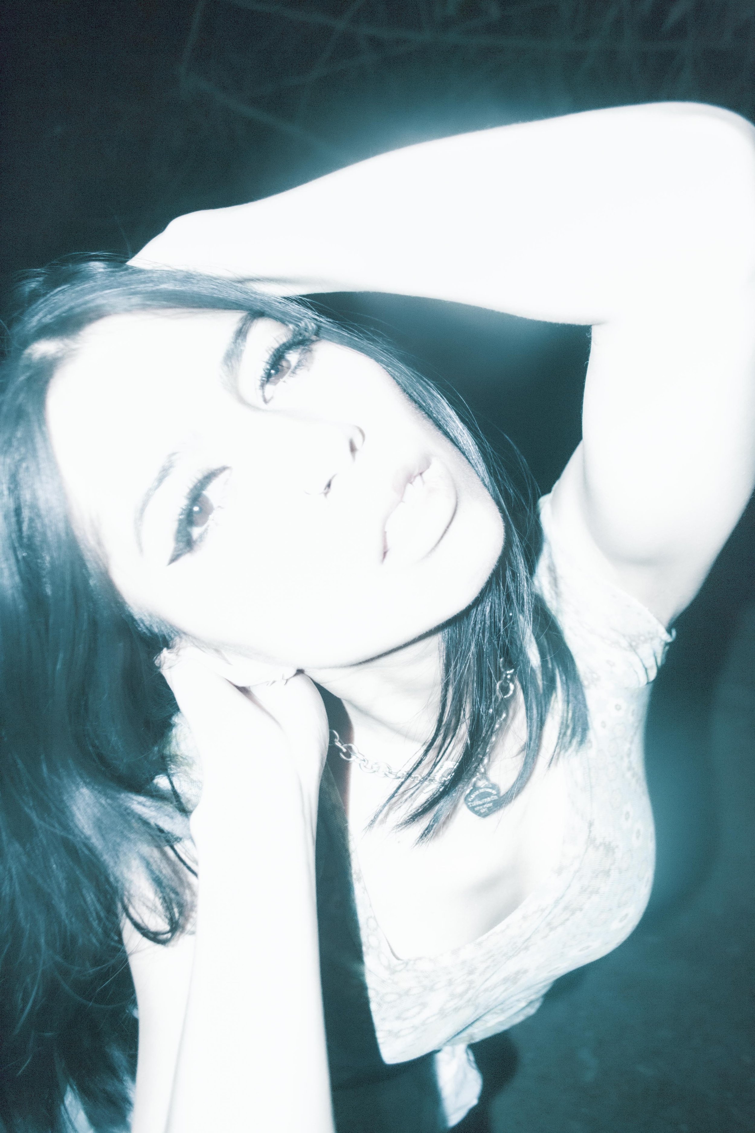 Black-haired woman with her head resting in her hand, looking into the distance with a soft expression, wearing a light-colored lace top and a necklace.