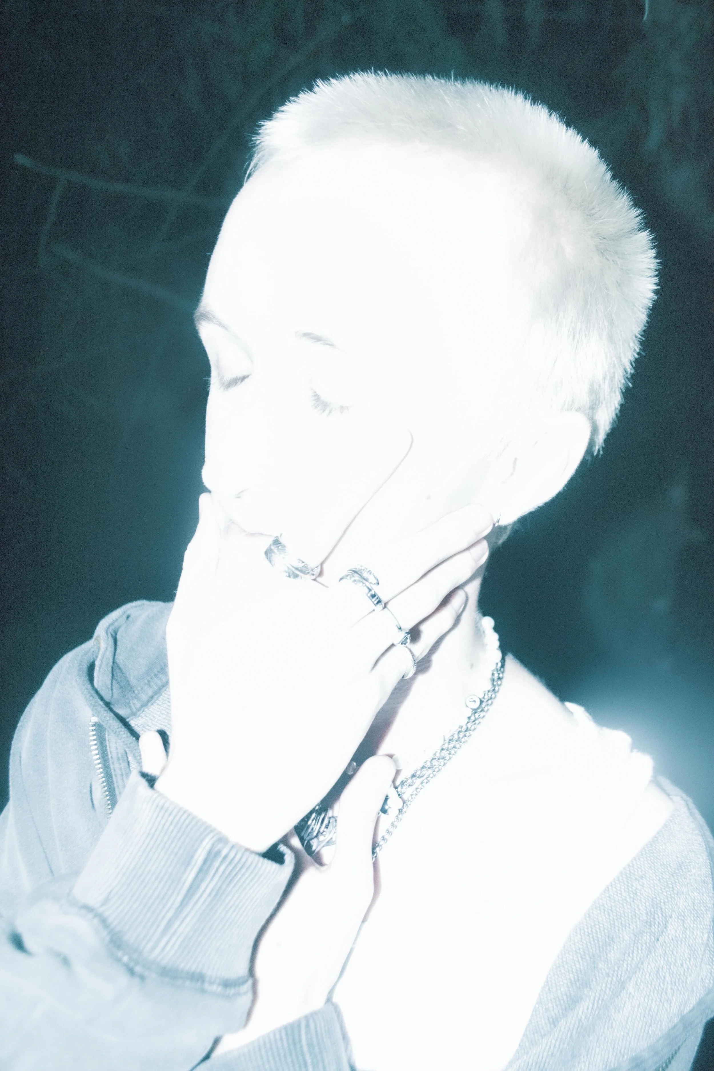 A person with short platinum blonde hair, light skin, and closed eyes, holding their hand to their neck on a dark background. They are wearing layered necklaces and jewelry, with a hoodie and multiple rings.
