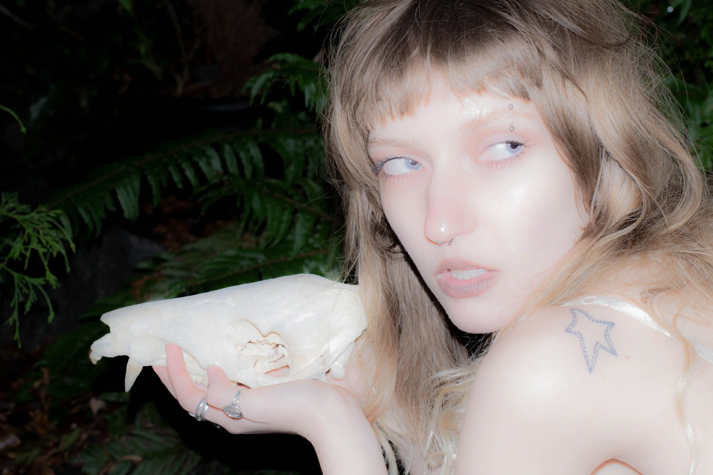 A young woman with light brown, wavy hair and facial piercings, holding a large piece of white coral or rock, standing outdoors with green foliage in the background.