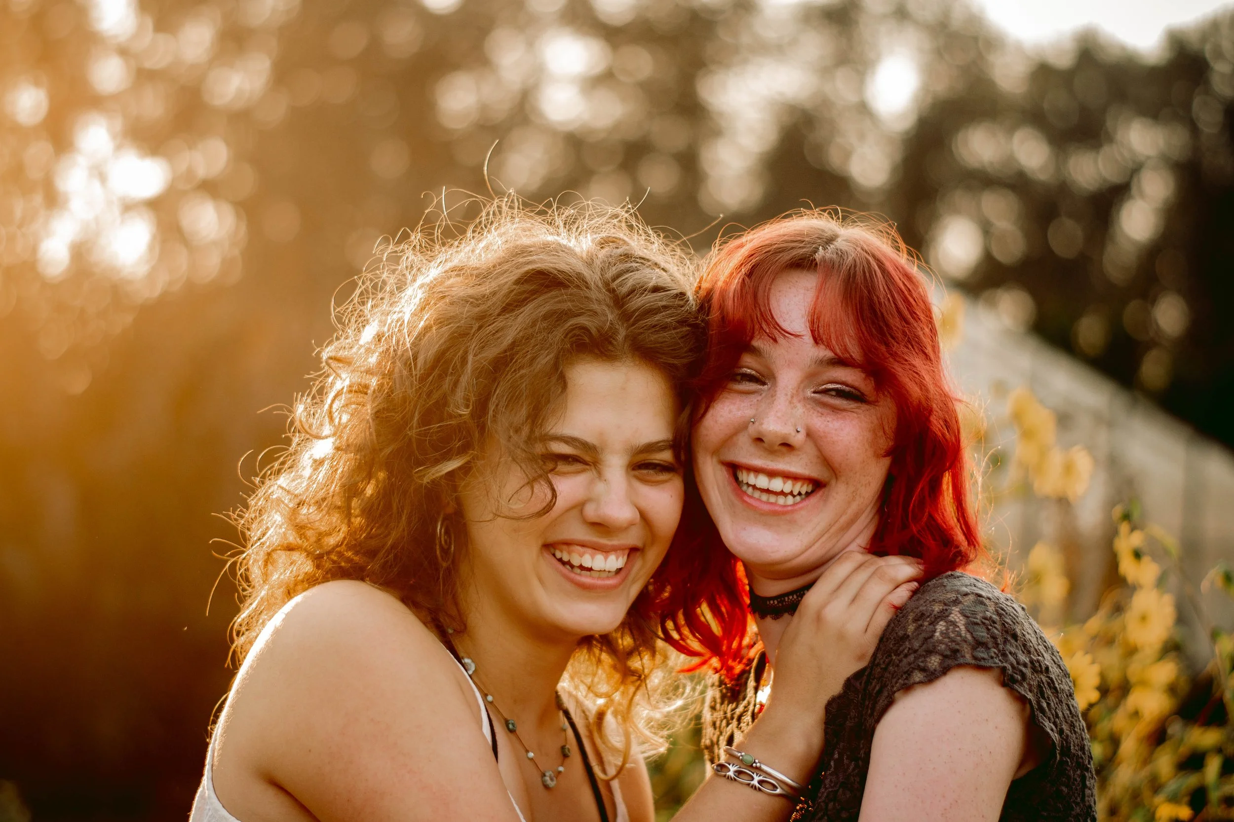 Two smiling women hugging outdoors with sunlight and blurred trees in the background.