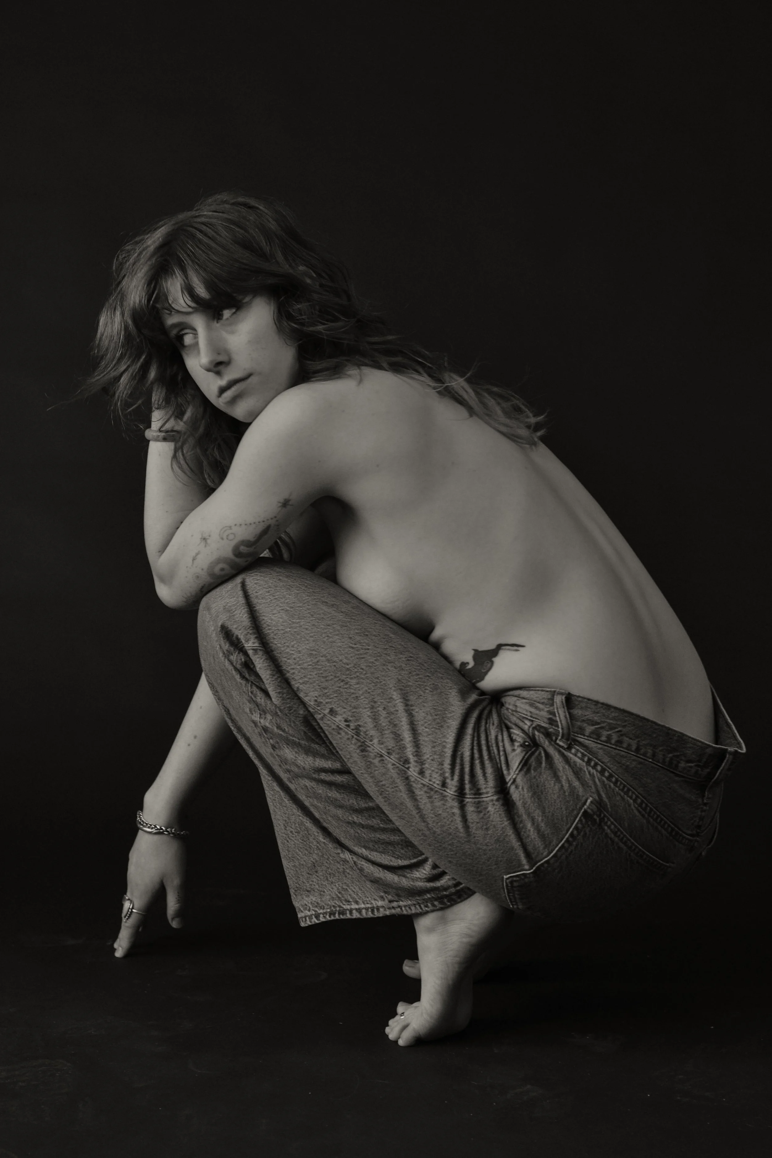 Black and white photo of a woman with tattoos, crouching with one hand on the ground and the other resting on her knee, wearing jeans, against a dark background.