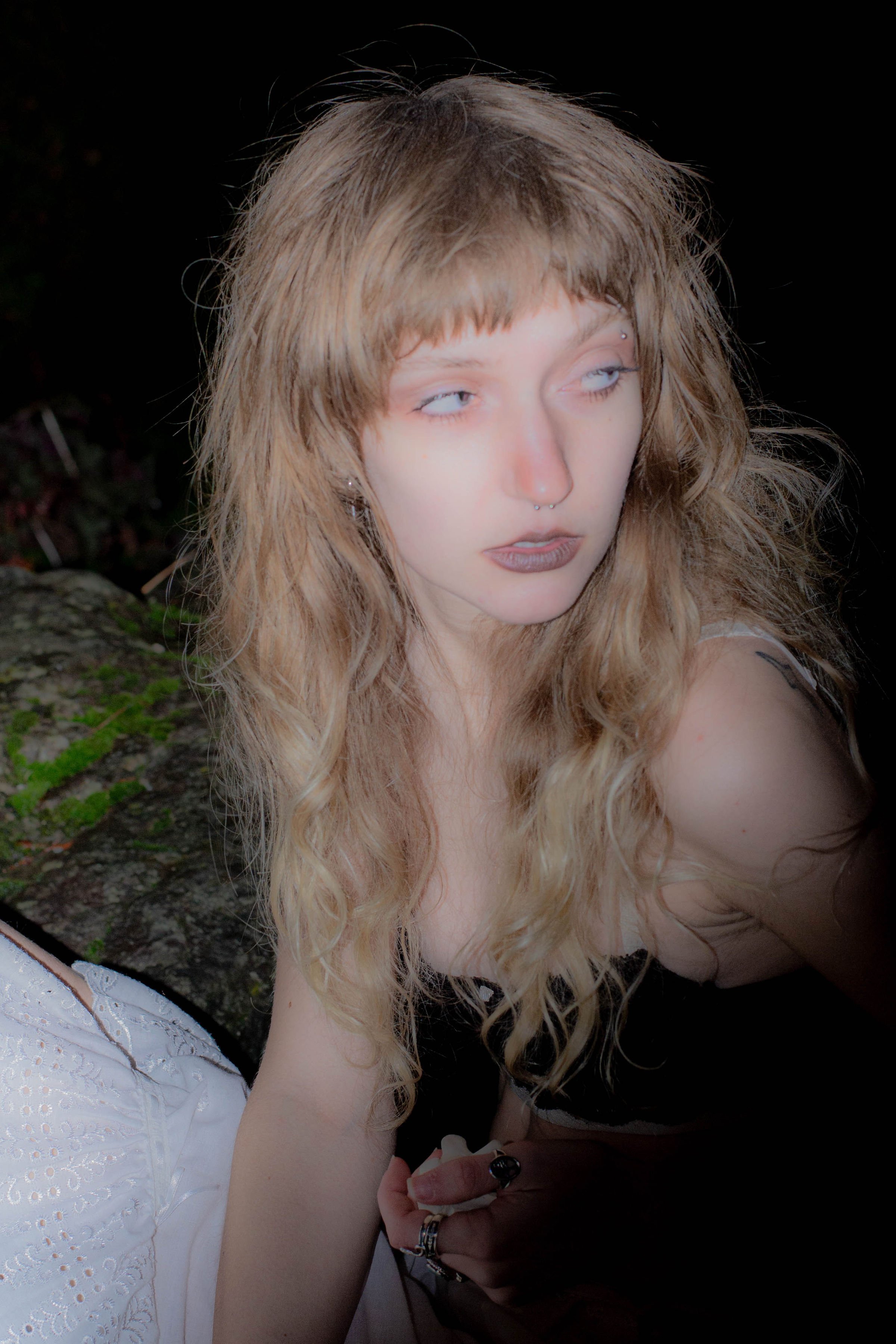 A young woman with long, wavy blonde hair and light makeup, including dark lipstick, is seen outdoors at night. She is looking away from the camera with a neutral expression and has visible facial piercings.