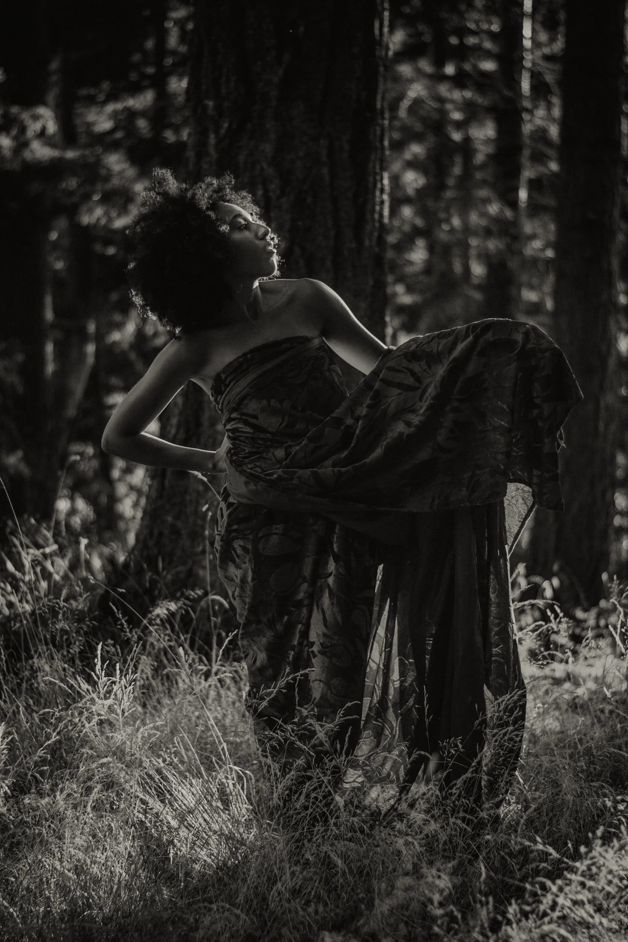 Black and white photo of a woman with natural curly hair sitting in a forest, wearing a strapless top and patterned skirt, looking contemplative.