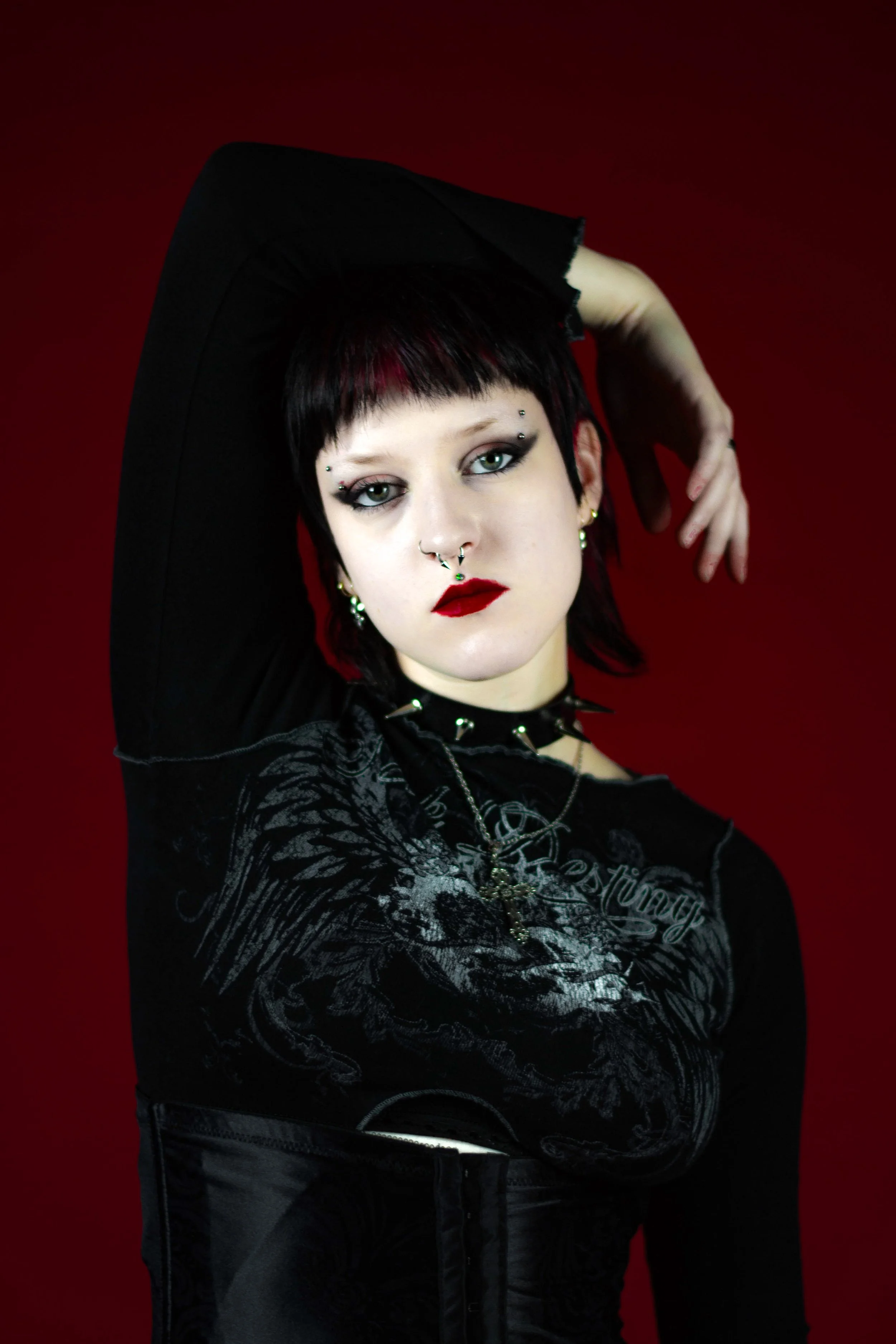 A person with short black hair and green eyes wearing makeup and piercings, dressed in dark clothing with gothic accessories, posed with one arm raised against a dark red background.