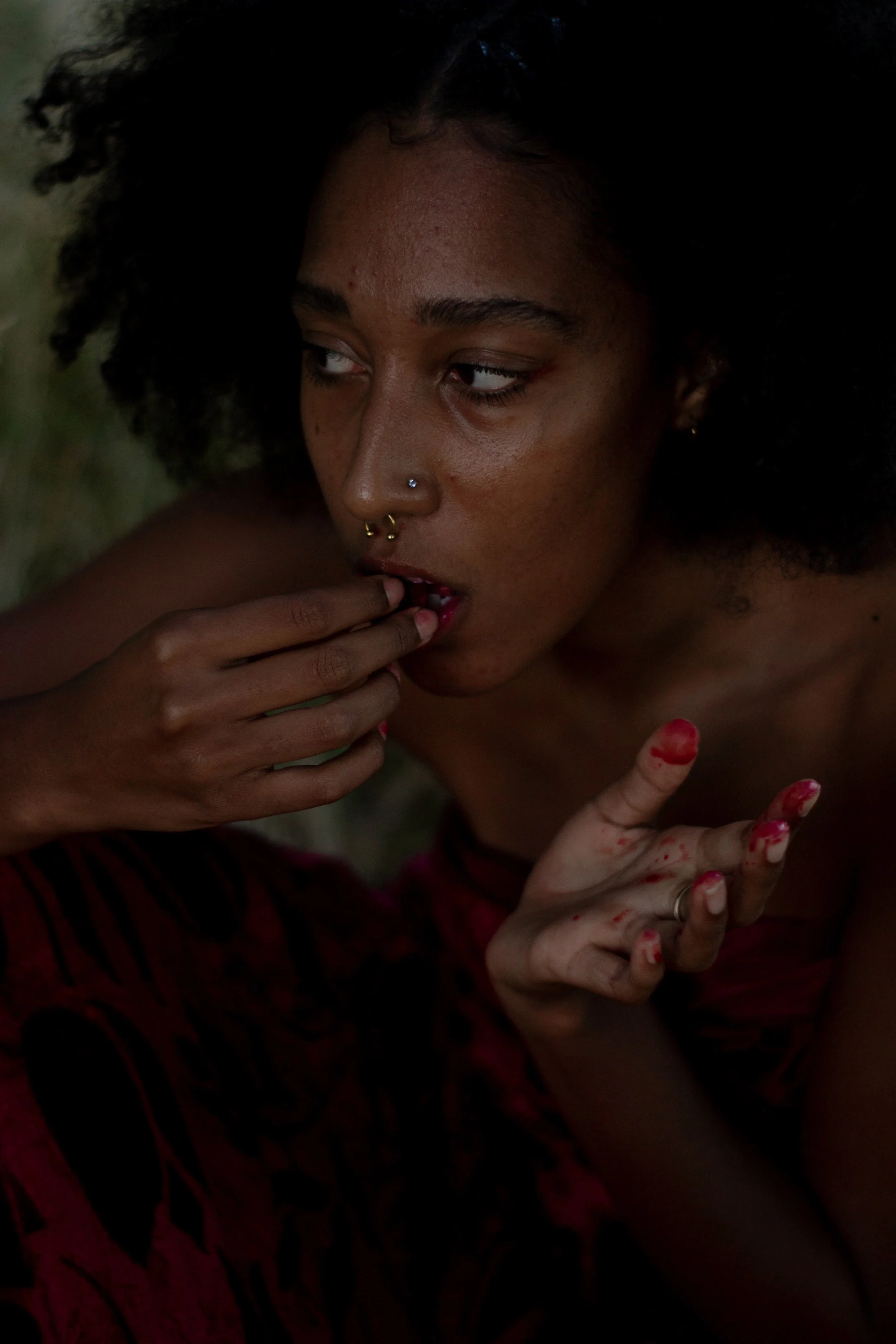 Close-up of a woman with dark, curly hair and multiple facial piercings, including a nose ring and septum piercing. She is touching her lips with her fingers, which are stained with red lipstick or possibly blood, and her eyes are looking to the side
