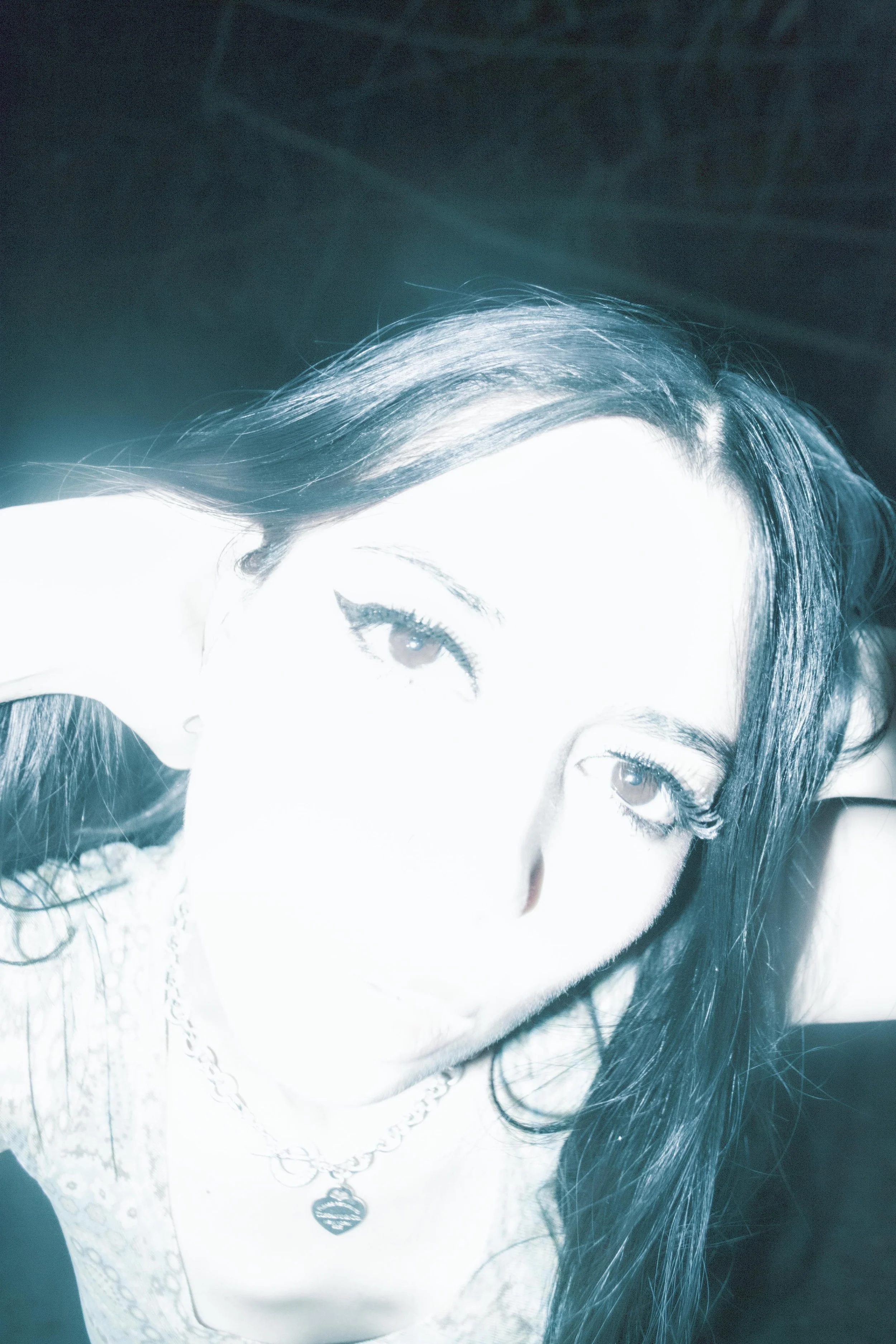 Close-up of a woman with dark hair and light skin, wearing necklaces, looking at the camera, with a dark background.