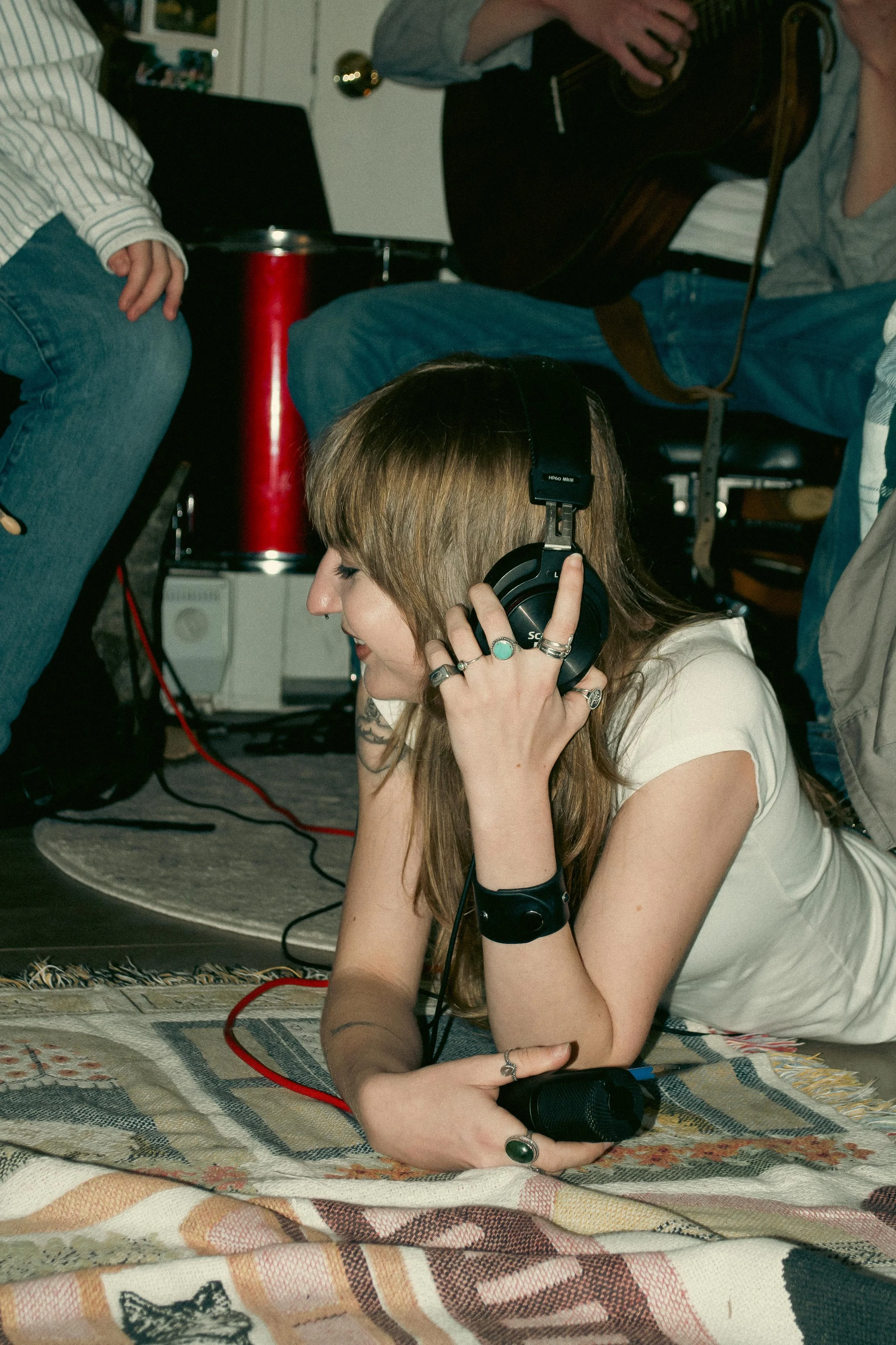 A woman with long light brown hair is sitting on the floor, wearing headphones, a white t-shirt, and silver jewelry. She is holding a black microphone in her right hand and appears to be smiling or singing. There are various objects around her, inclu