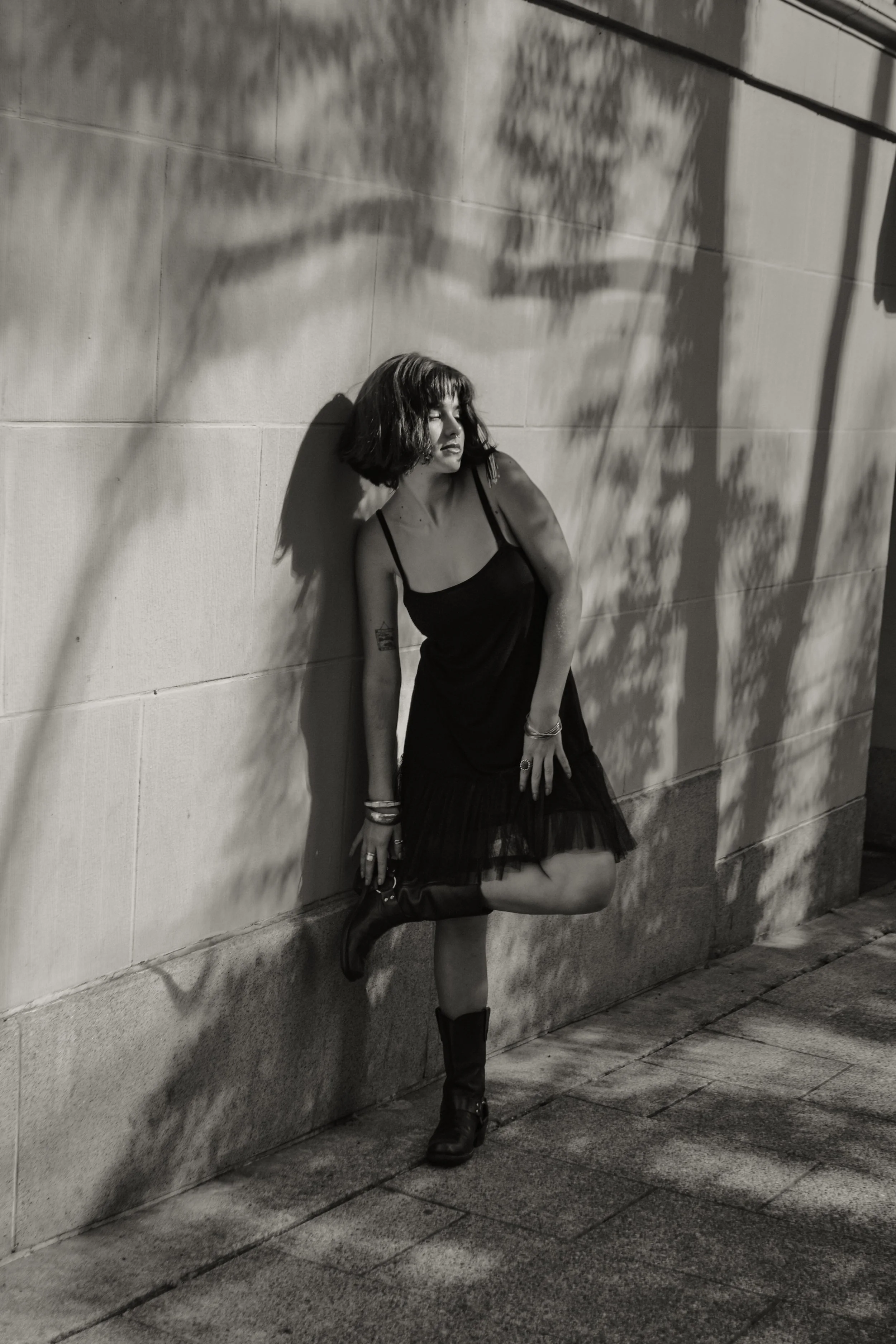 A woman leaning against a concrete wall, casting shadows of tree branches on the wall, in black and white. She is dressed in a black dress, wearing black boots, and has short, wavy hair.