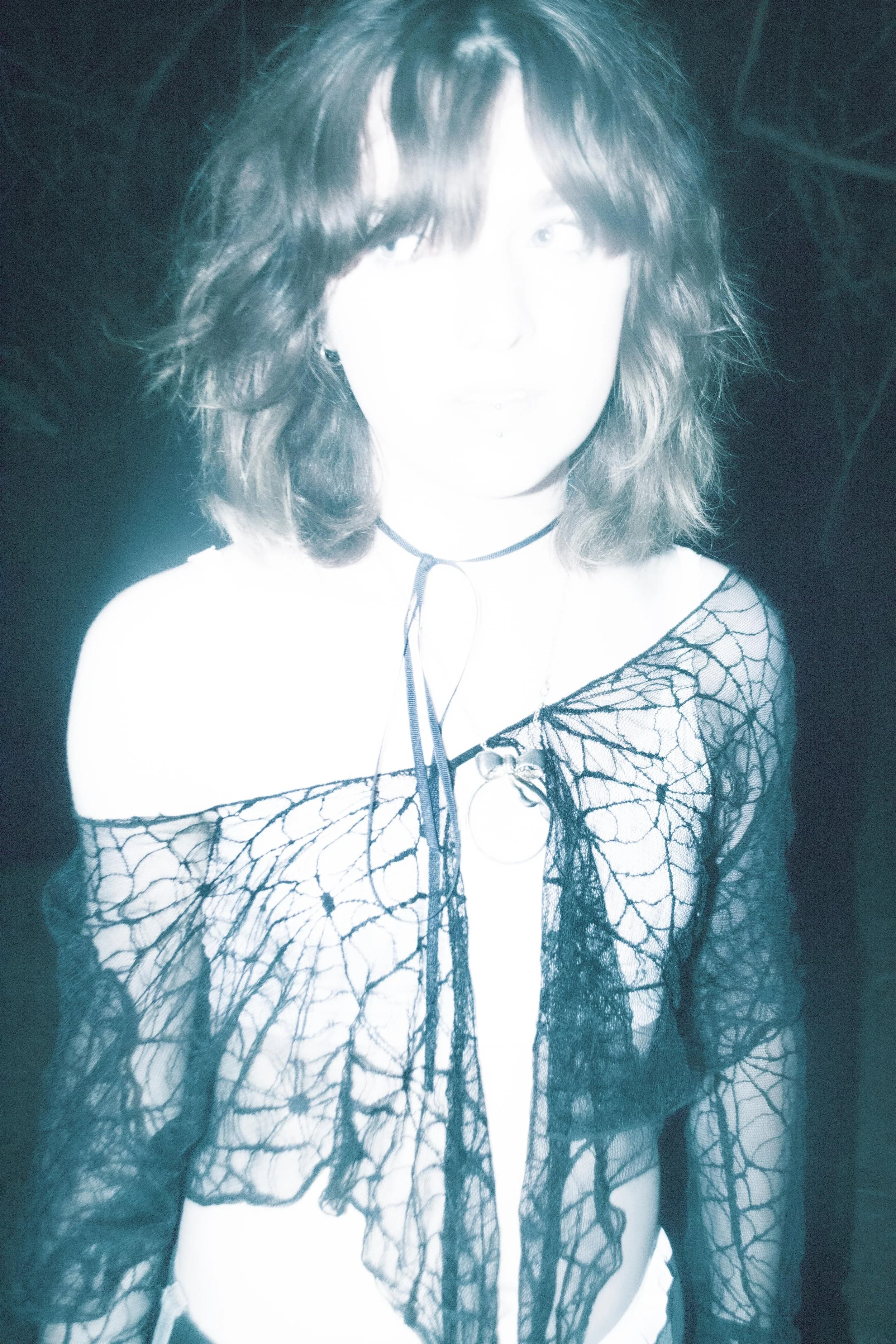 A person with shoulder-length hair wearing a black netted top with a spiderweb pattern, posing against a dark background.