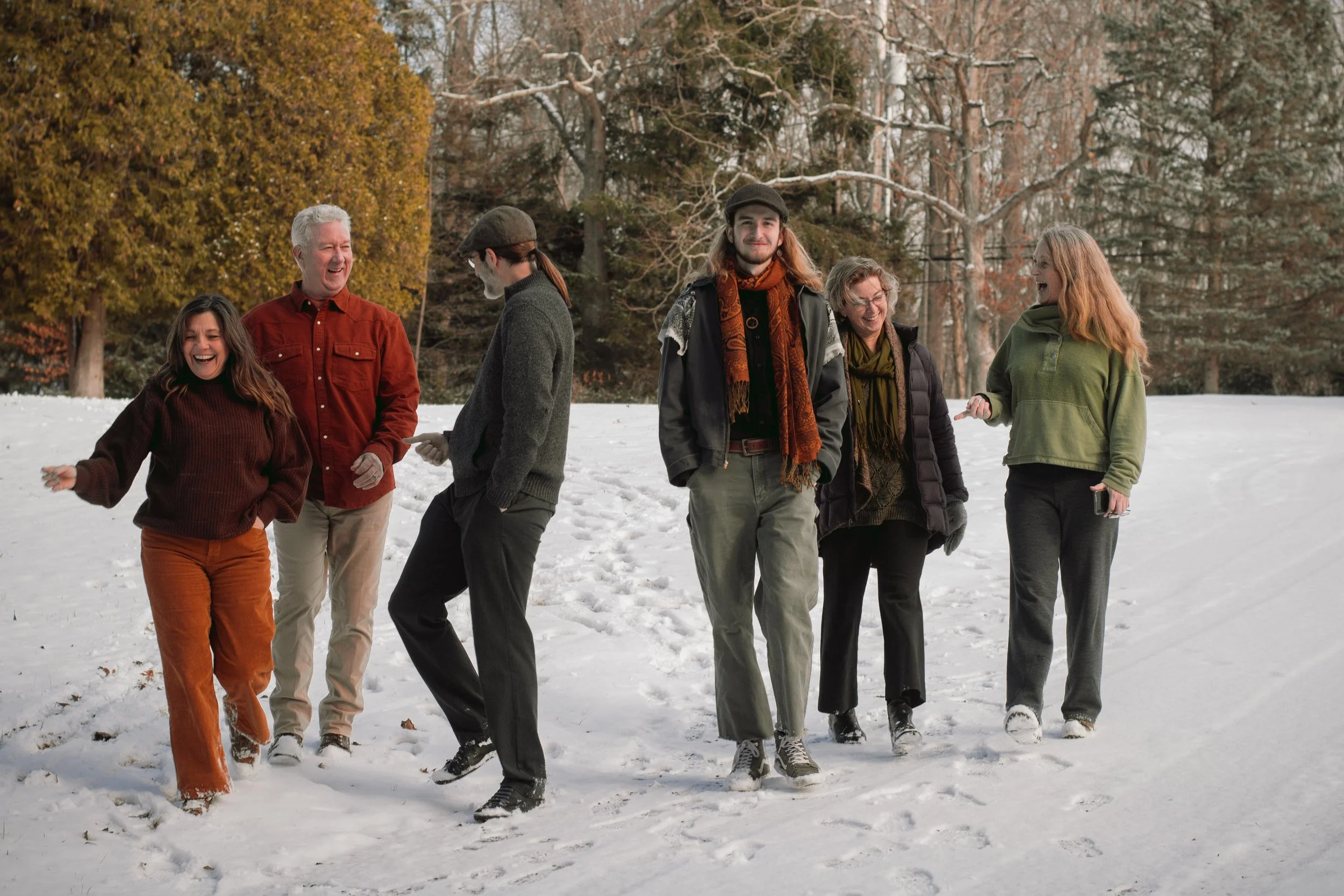 Group of six people walking in snow-covered park surrounded by trees, laughing and enjoying winter weather.