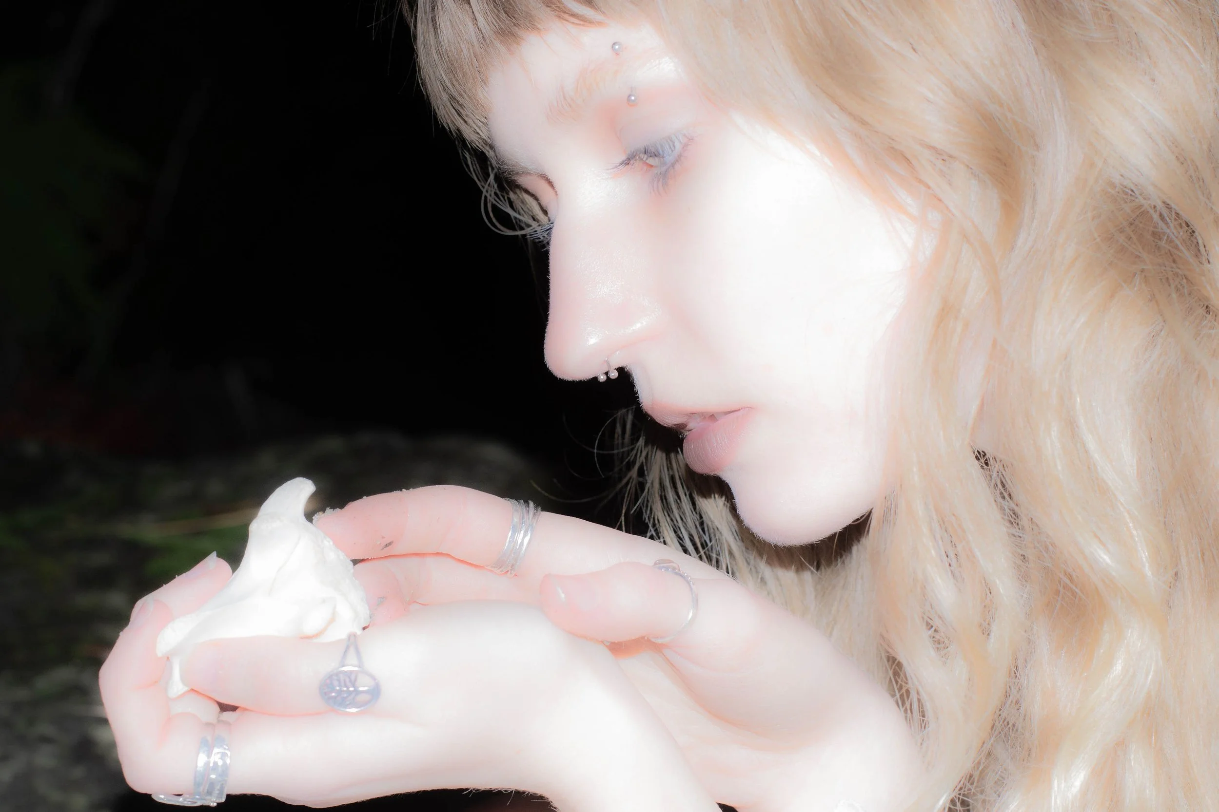 A close-up of a woman with light-colored hair, Piercings on her eyebrow, nose, lips, and ears, holding a dollop of cream or lotion in her hand.