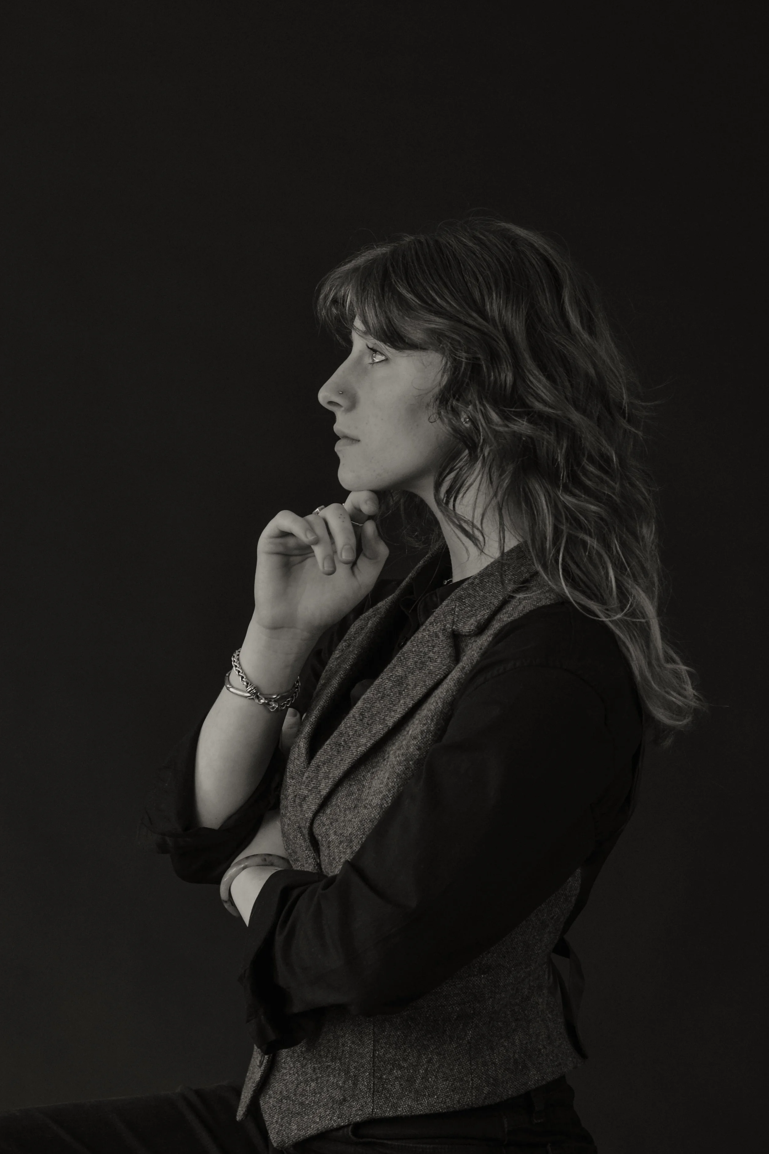 A black and white photo of a woman in profile. She has wavy hair, a nose piercing, and is wearing a blazer. She is touching her chin with her hand, looking contemplative against a dark background.