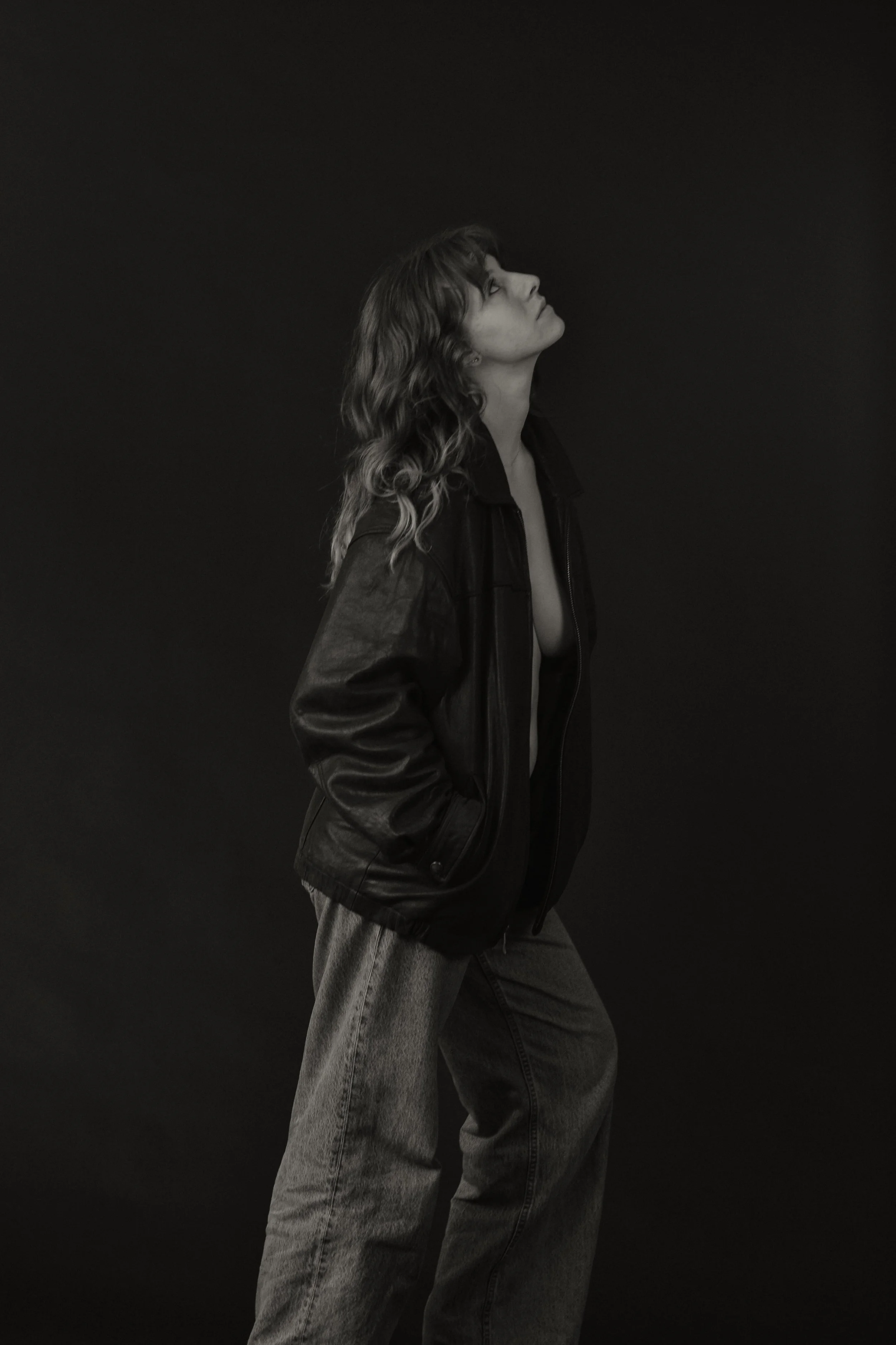 A young woman with wavy hair wearing a black leather jacket and baggy jeans stands against a dark background, looking upward with her lips slightly pursed.
