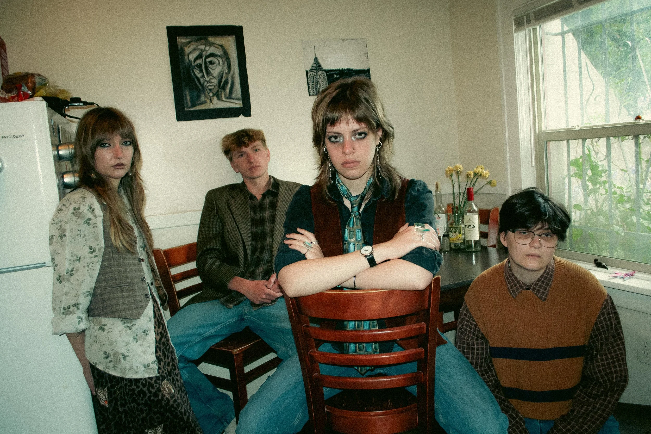 Five young people at a dining table, two women and three men, in a room with artwork on the wall and a window, some bottles on the table.