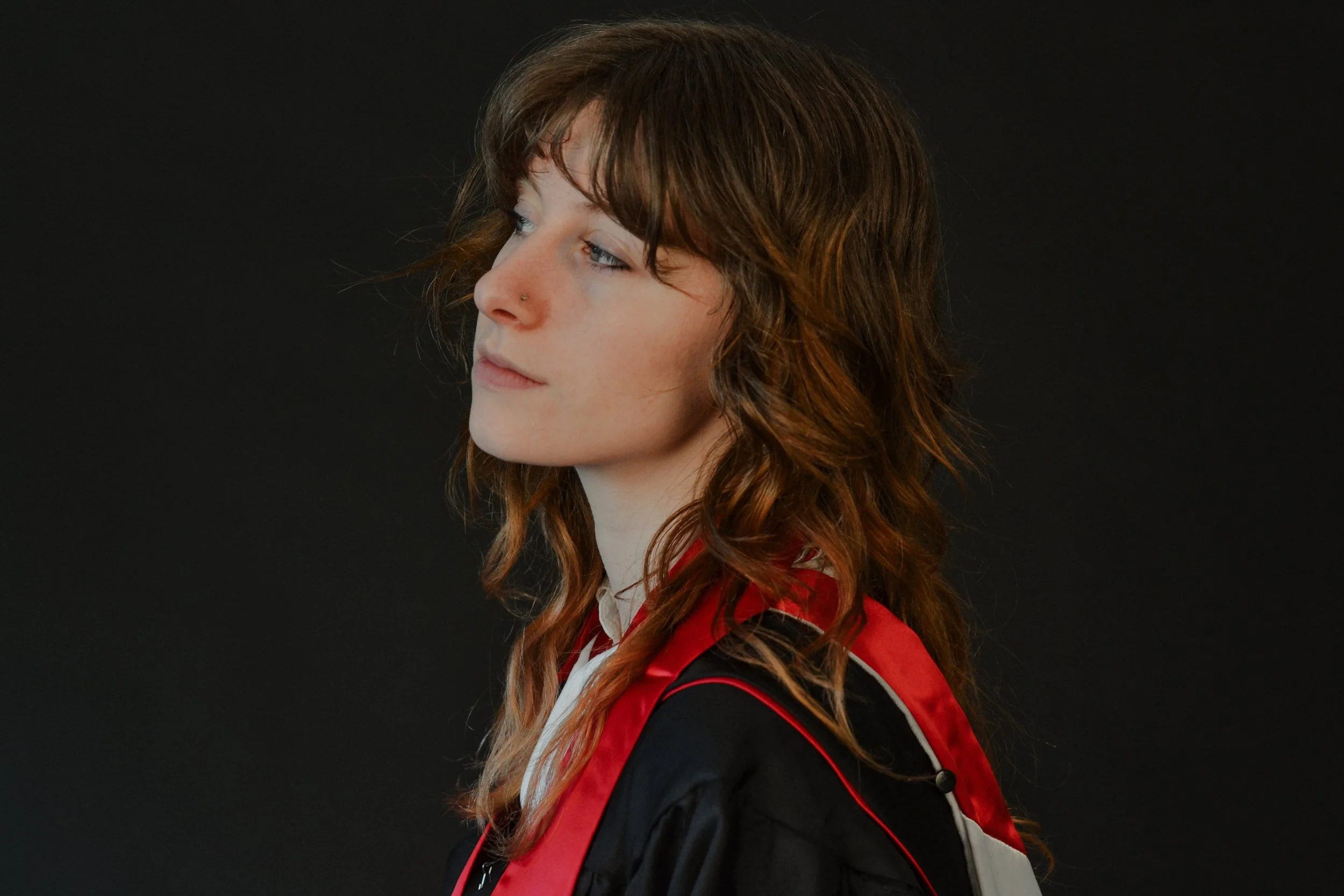Profile of a young woman with wavy auburn hair, wearing a red and black jacket, against a dark background.
