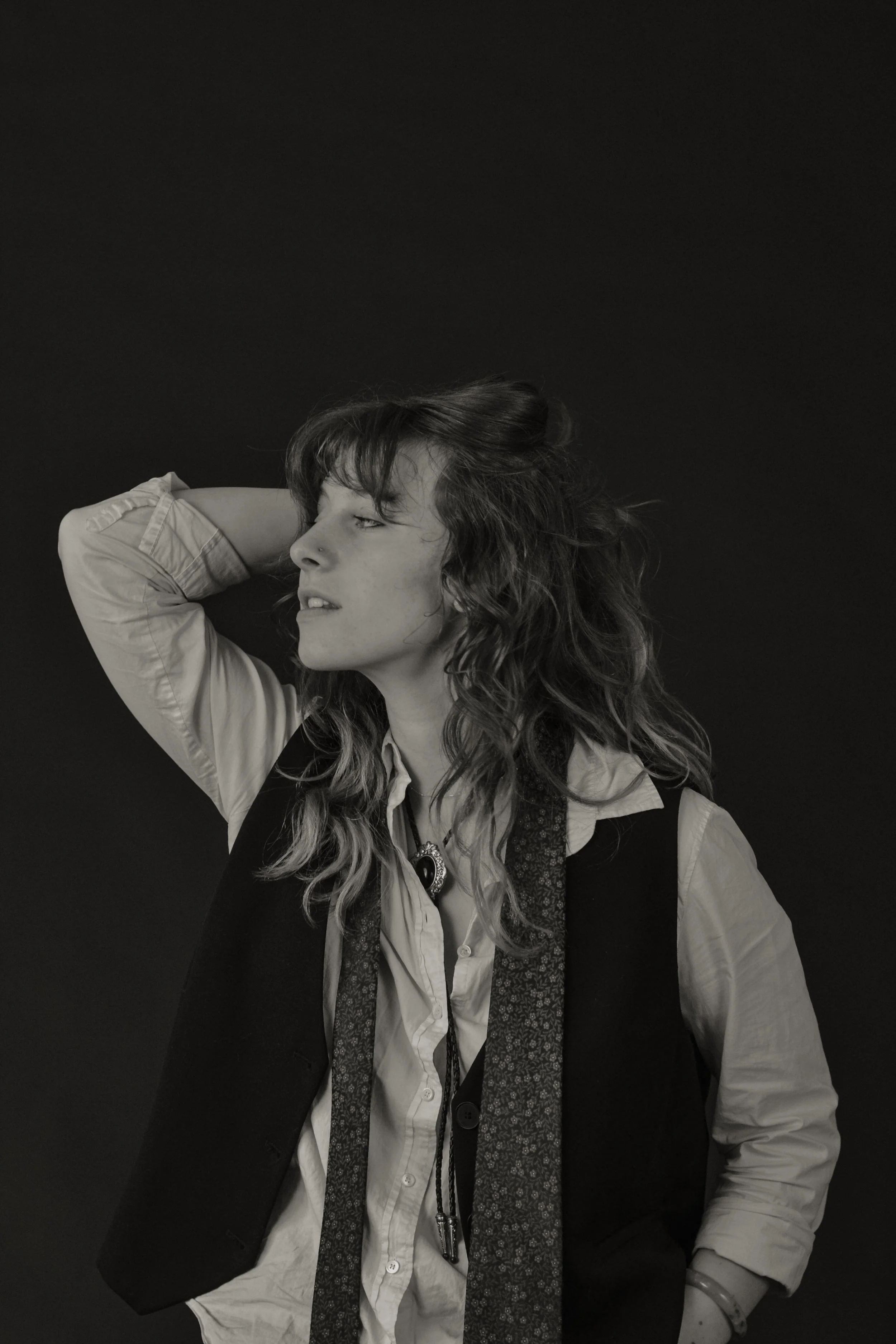 Black and white photo of a woman with curly hair, wearing a shirt, vest, and layered jewelry, striking a pose with one arm raised and hand resting behind her head, against a dark background.