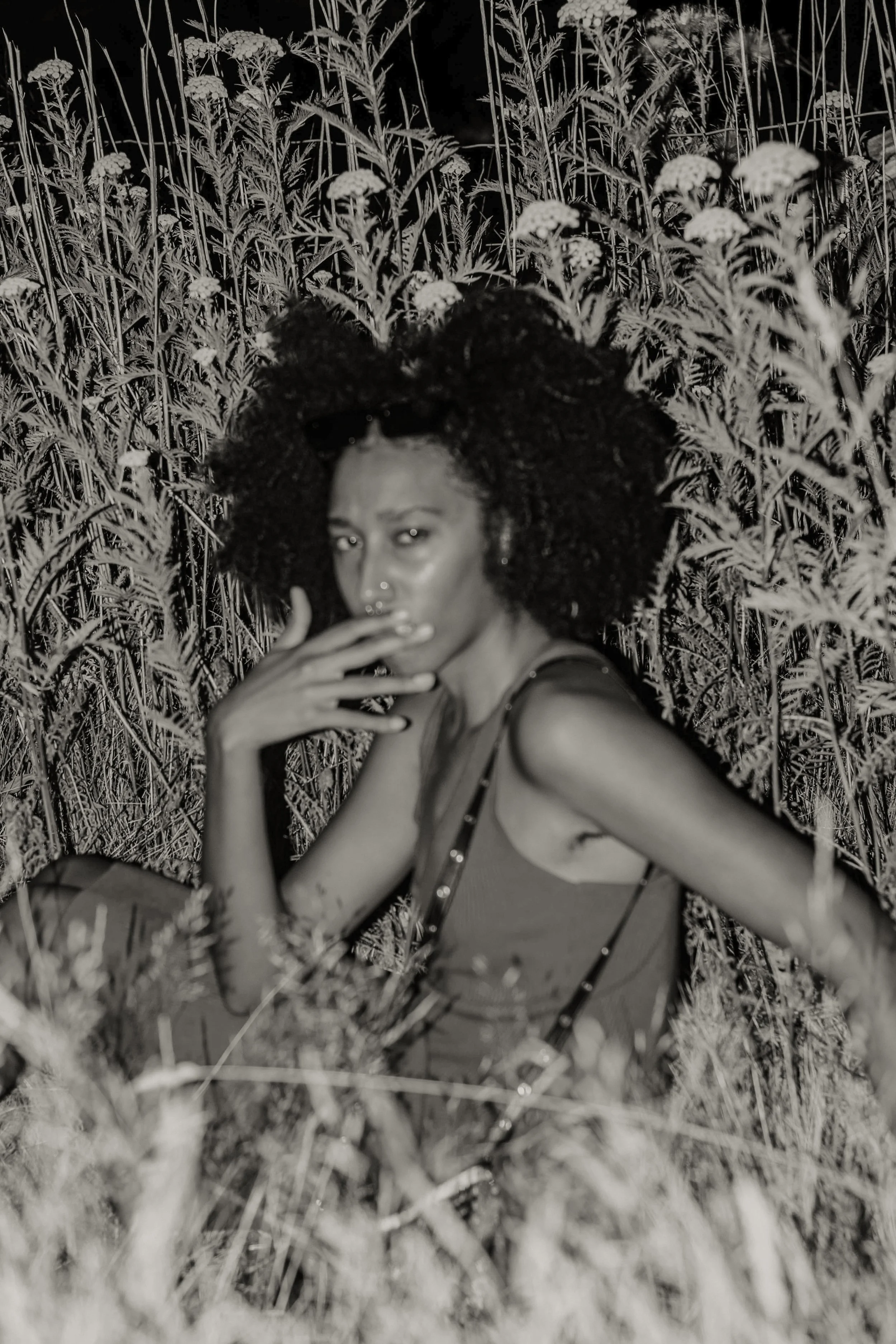 A woman with curly hair, wearing a tank top, taking a selfie in a field of tall wildflowers at night.