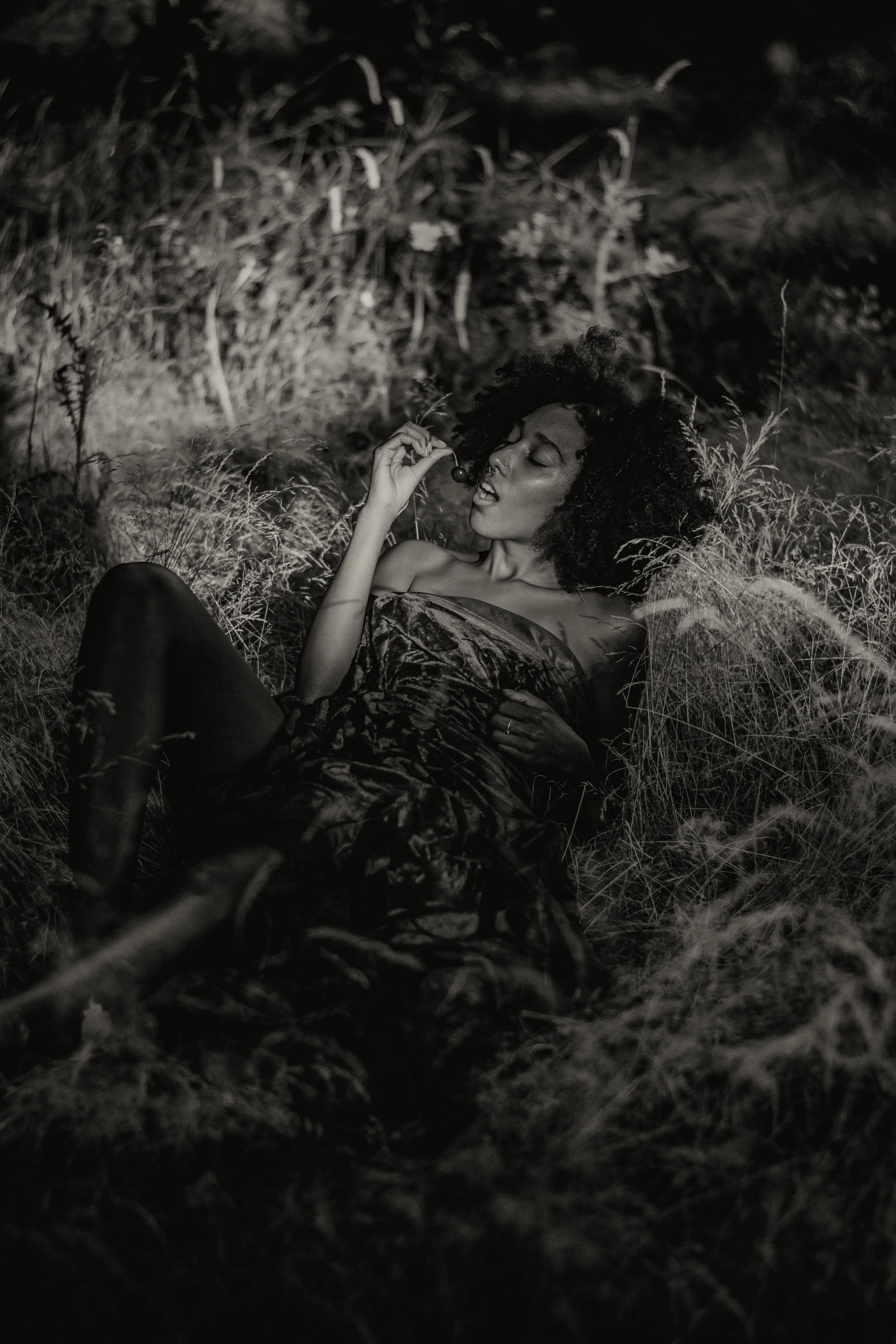 A woman with curly hair lying in grass, holding a cherry near her mouth, in black and white.
