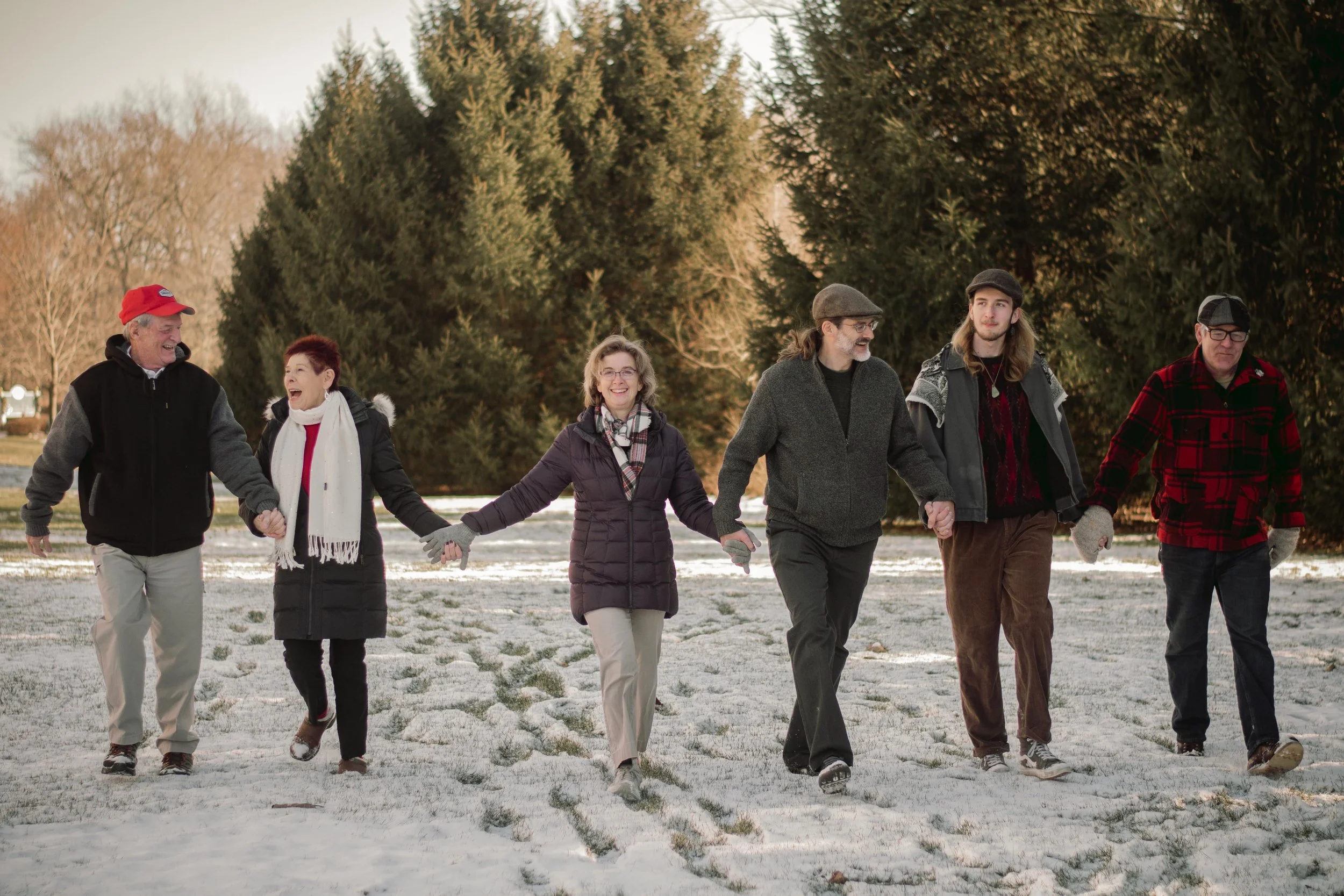 A group of six people, holding hands, walking on a snowy field with evergreen trees in the background. They are dressed in winter clothing and appear to be enjoying a walk outdoors.