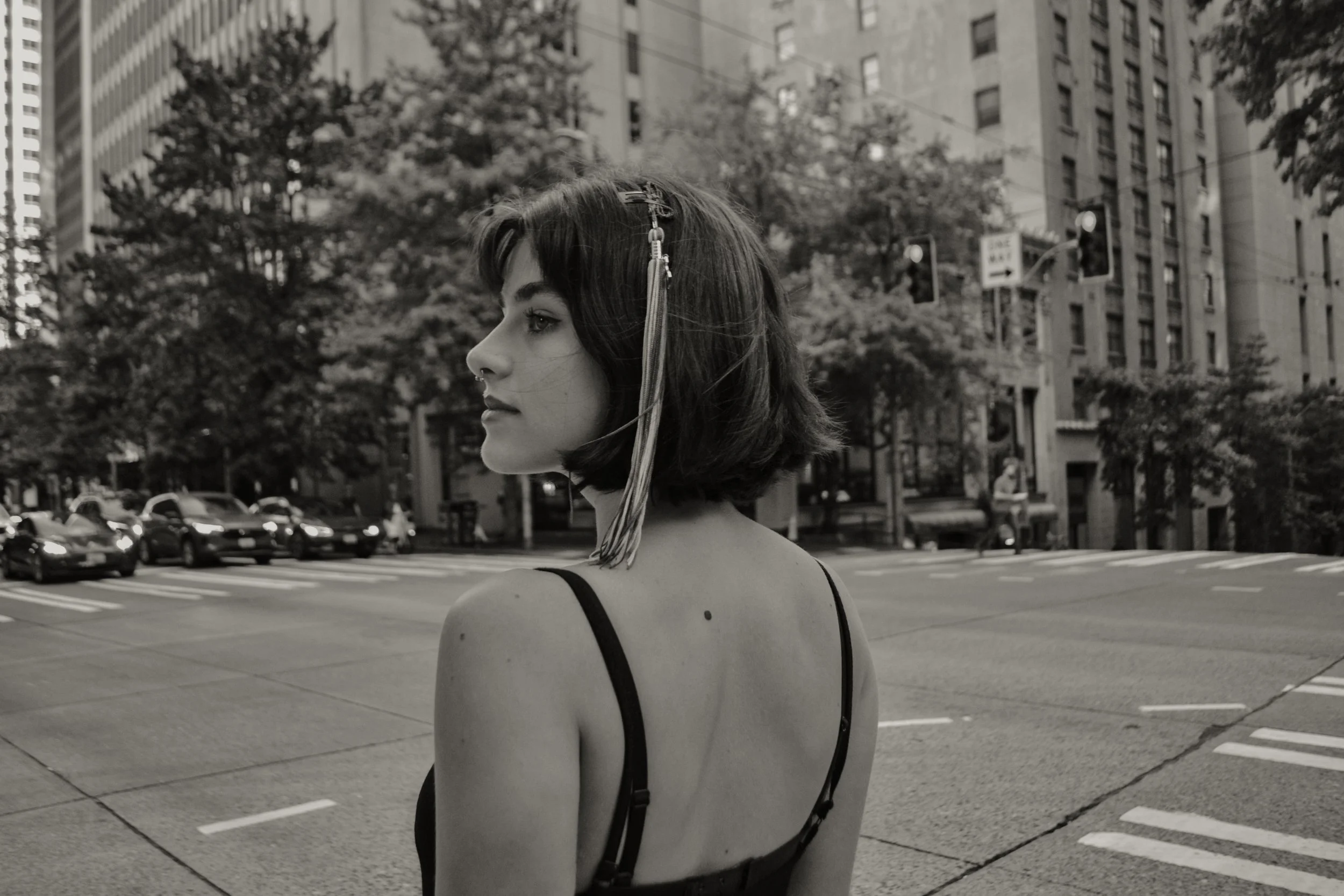 A young woman with shoulder-length dark hair wearing a tank top and a clip in her hair stands on a city street corner. She is looking to her left, with a serious expression, in front of tall buildings. There are cars and trees visible in the background, and the photo is in black and white.