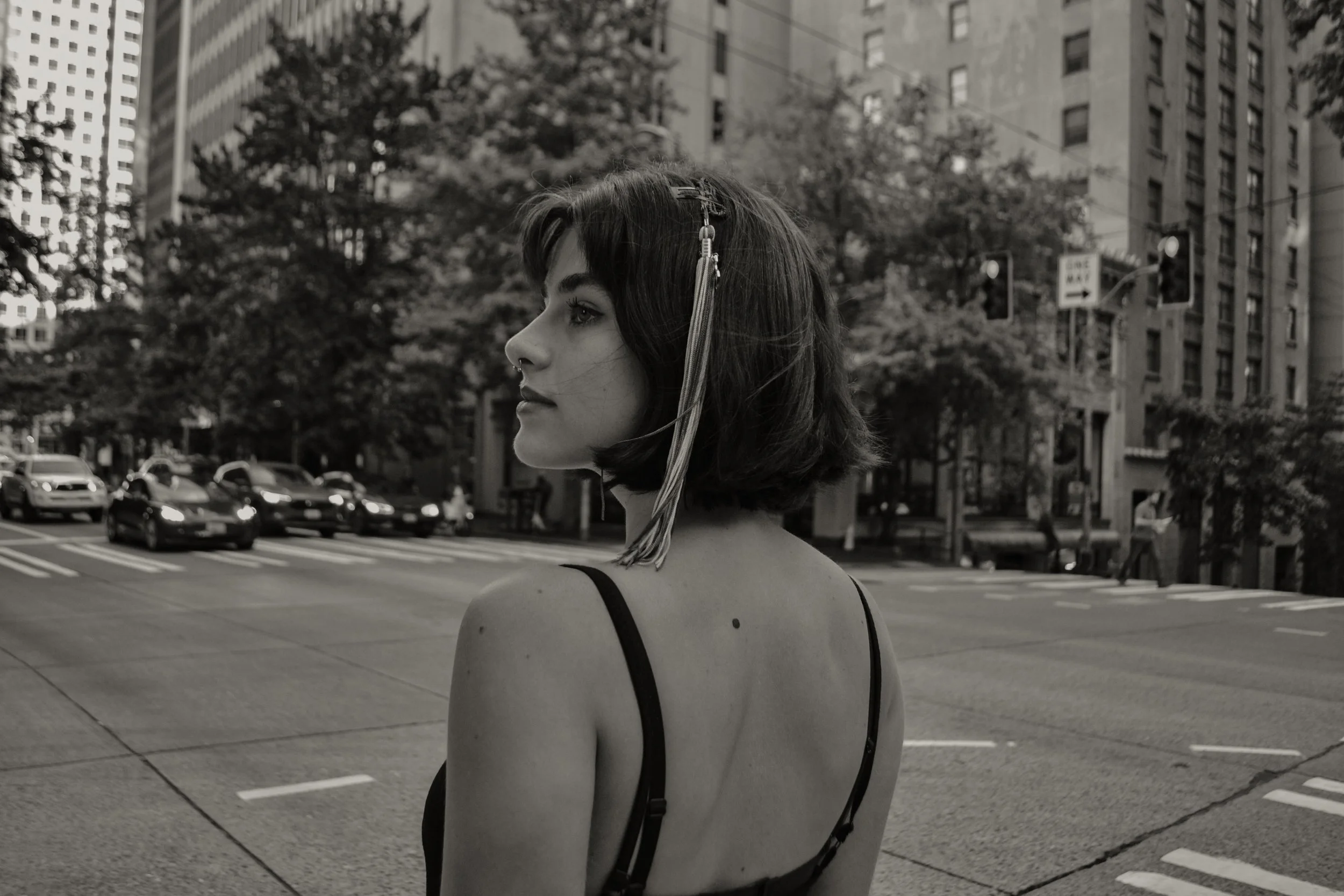 A young woman with dark hair, a piercing, and a decorative hairpin stands in an urban city street. The background features tall buildings, a street sign, trees, and parked cars. The photo is in black and white.