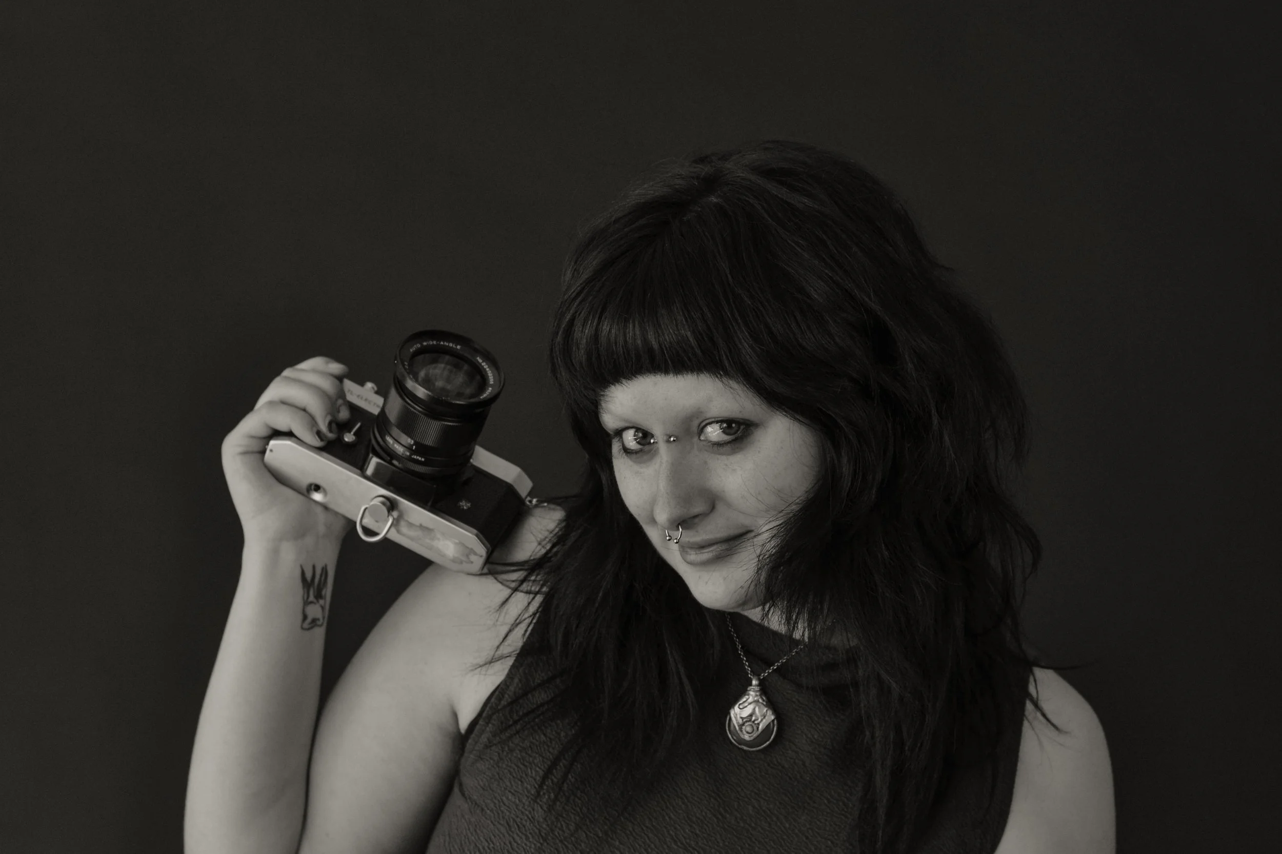 Black and white photo of a woman with dark hair and bangs, holding a vintage camera up to her face, looking over it, wearing a necklace and a dark tank top.