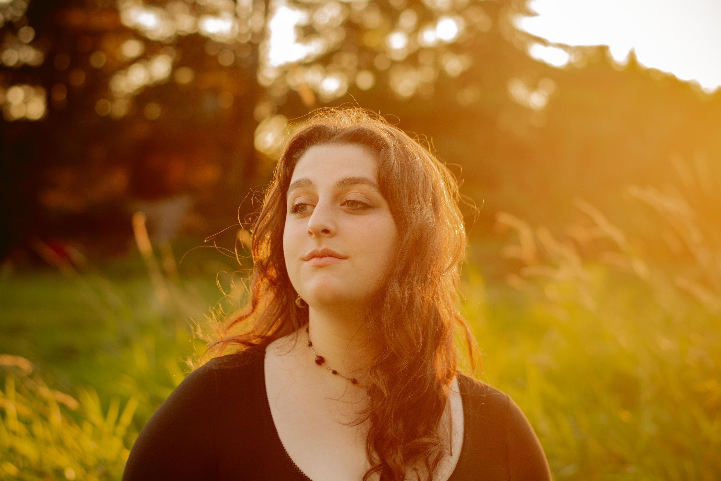 A young woman with long, wavy red hair standing outdoors during sunset with golden light illuminating her face and hair, surrounded by blurred greenery and trees.