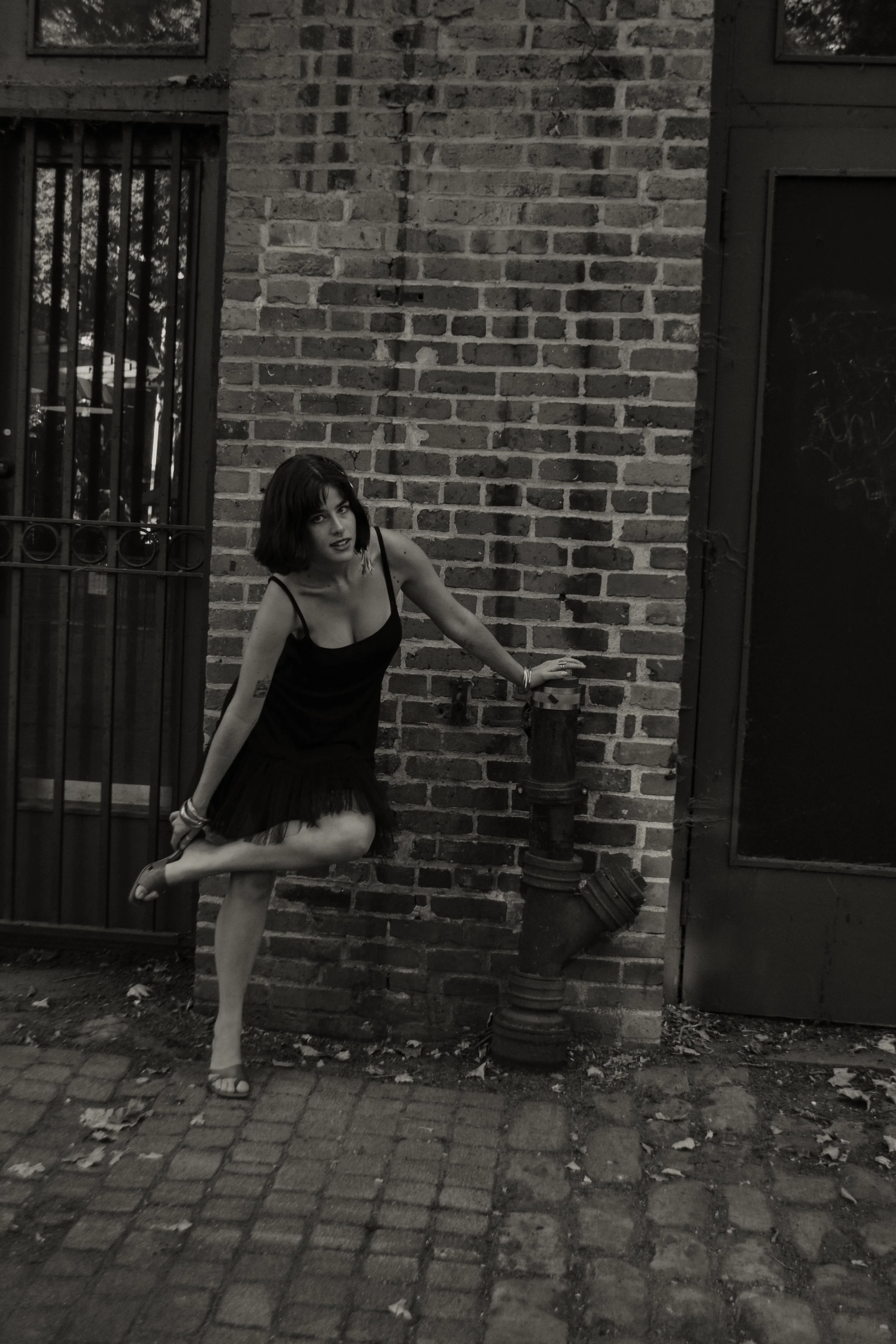 A woman in a black dress and heels standing outdoors on a brick sidewalk with a brick wall and a metal gate behind her. She has dark, shoulder-length hair and is posing with one leg lifted, holding onto a post.