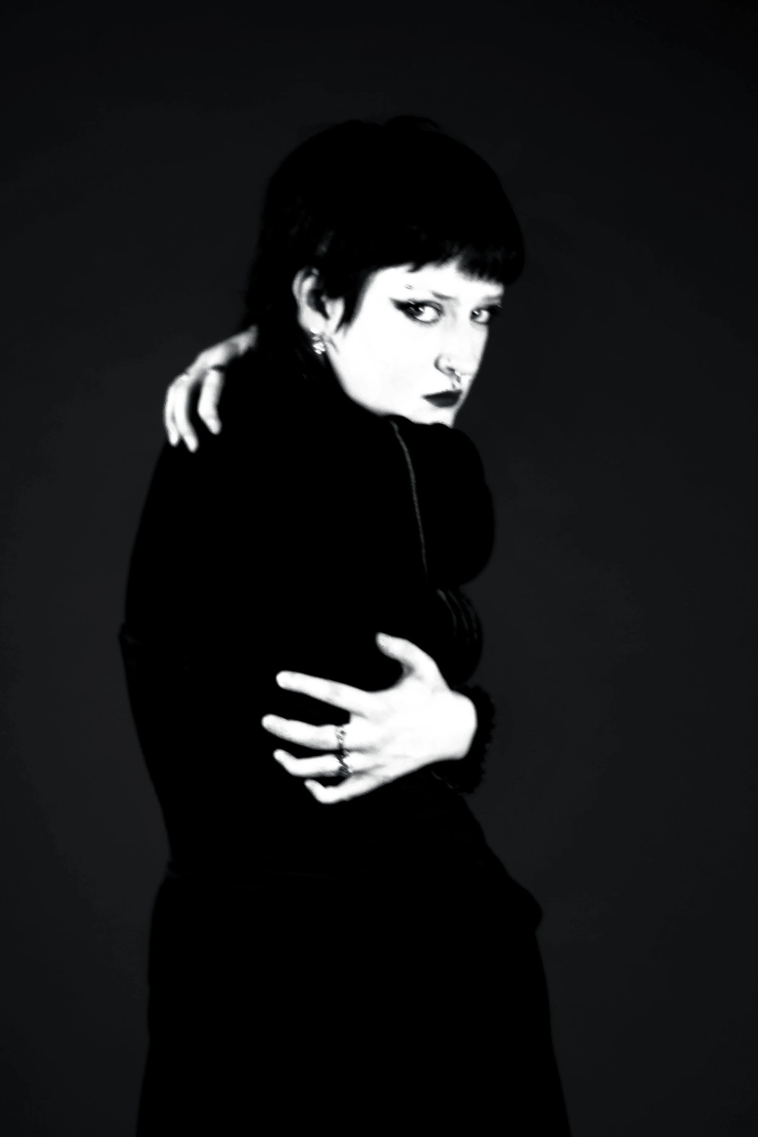 A black-and-white portrait of a woman with short dark hair, wearing dark makeup, and a dark outfit. She is holding herself with her arms crossed, with one hand resting on her shoulder and the other on her waist, looking intensely at the camera agains