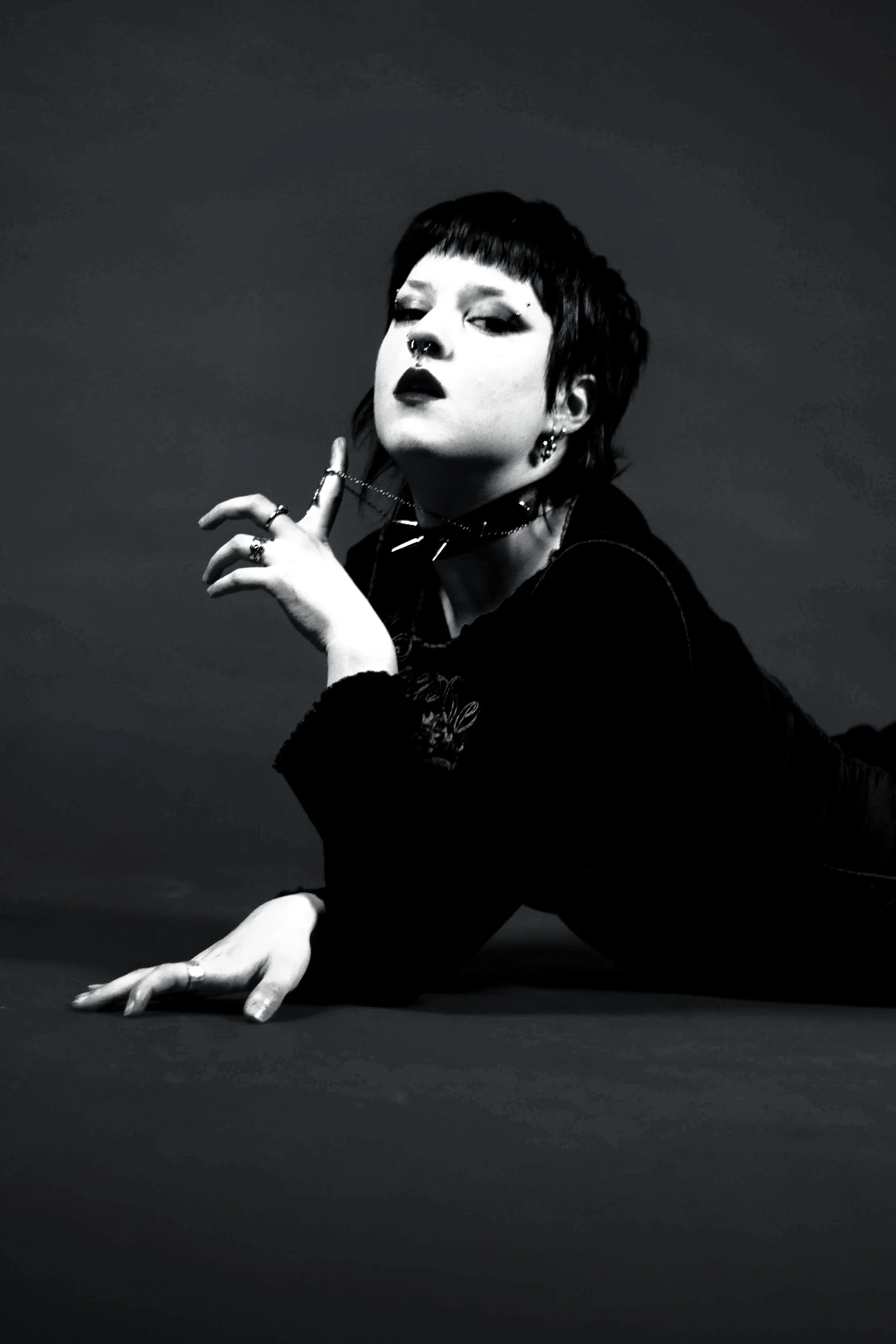 Black and white photo of a woman with short dark hair, dark lipstick, and piercings, posing with her hand near her face and her elbow on the ground against a plain dark background.