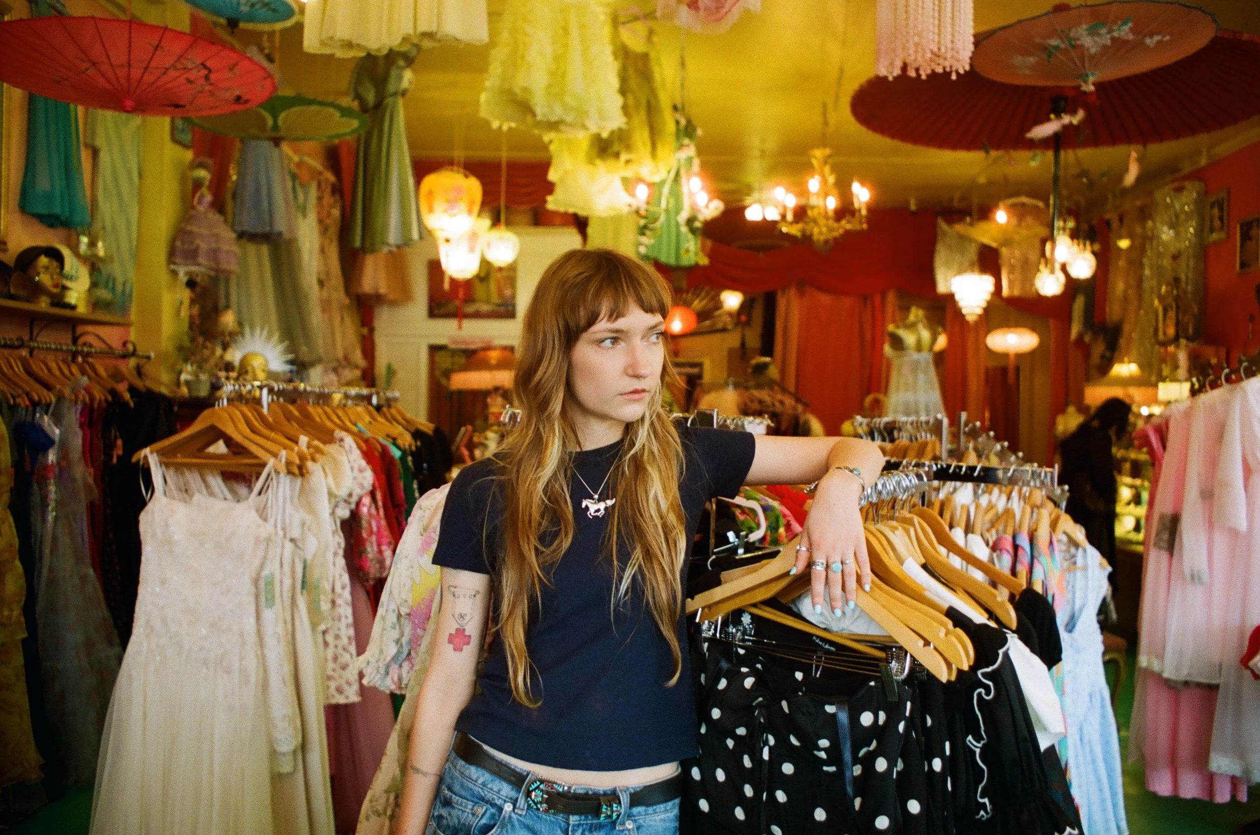 A young woman with long, light brown hair and tattoos on her arm is shopping for clothes in a vintage clothing store. She is wearing a black T-shirt with a necklaces and rings. She is holding clothes hanging on a rack and looking to her left.