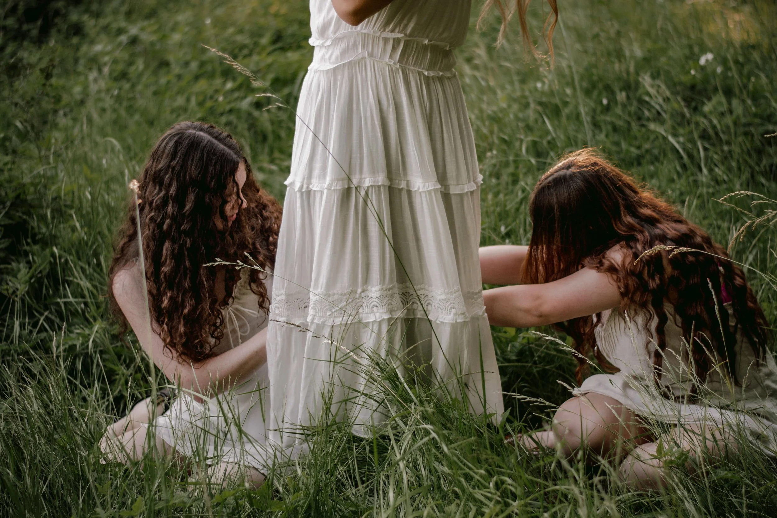 Three women with long, curly hair dressed in cream-colored dresses are sitting and kneeling in a grassy field, surrounding a white dress on a dummy, possibly during a photoshoot or costume fitting.