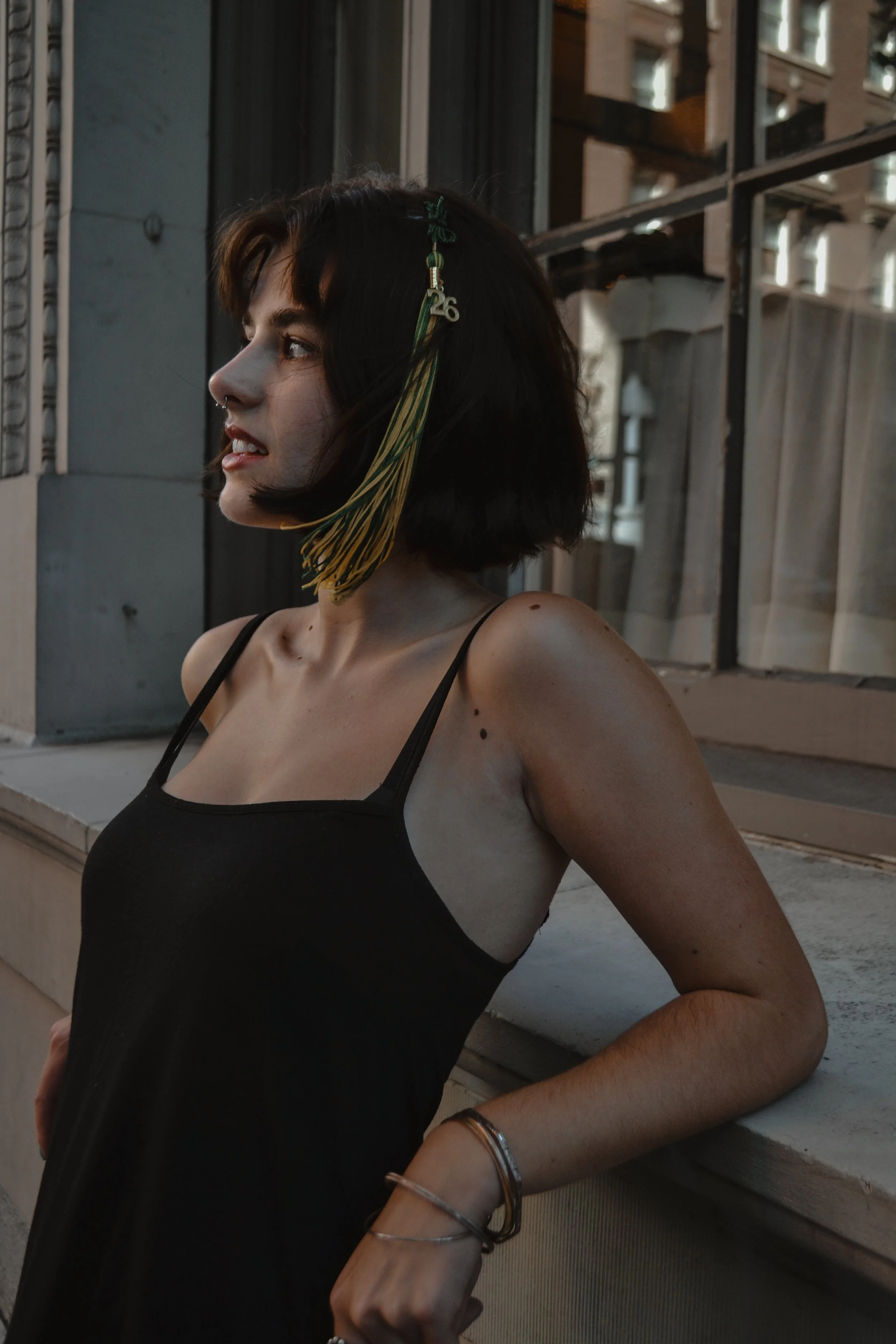 A woman wearing a black dress with thin straps stands outdoors near a building with large windows. She has short, dark hair with a hair accessory that features green and yellow ribbons and a metallic charm with the number 26. She is turned to the sid