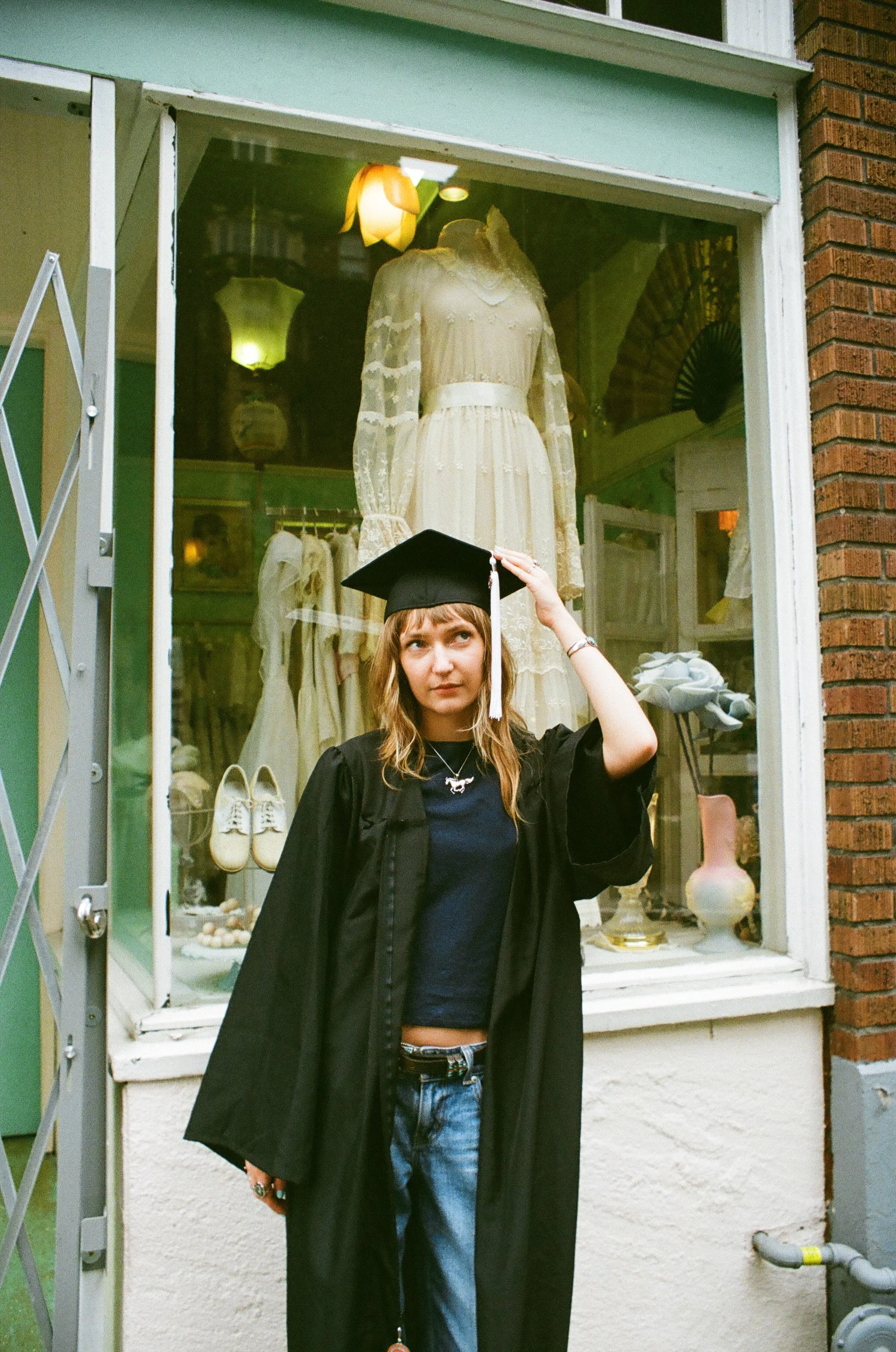 A young woman wearing a graduation cap and gown standing outside a storefront window with a vintage dress on display. She has light brown hair, blue eyes, and is wearing a navy blue shirt, jeans, and jewelry.