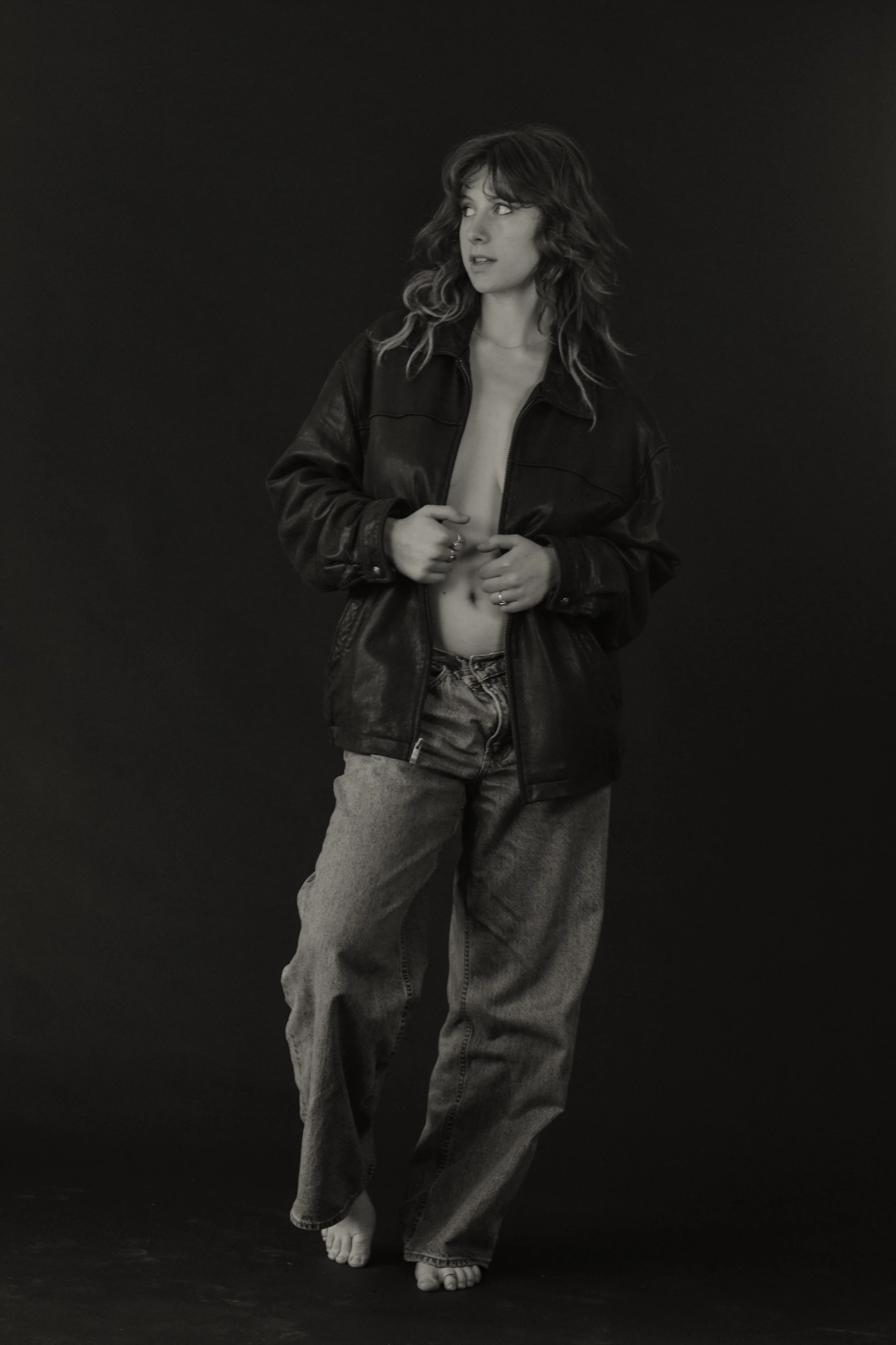 A black and white photo of a woman with wavy hair, wearing a leather jacket and high-waisted jeans, standing barefoot against a dark background.