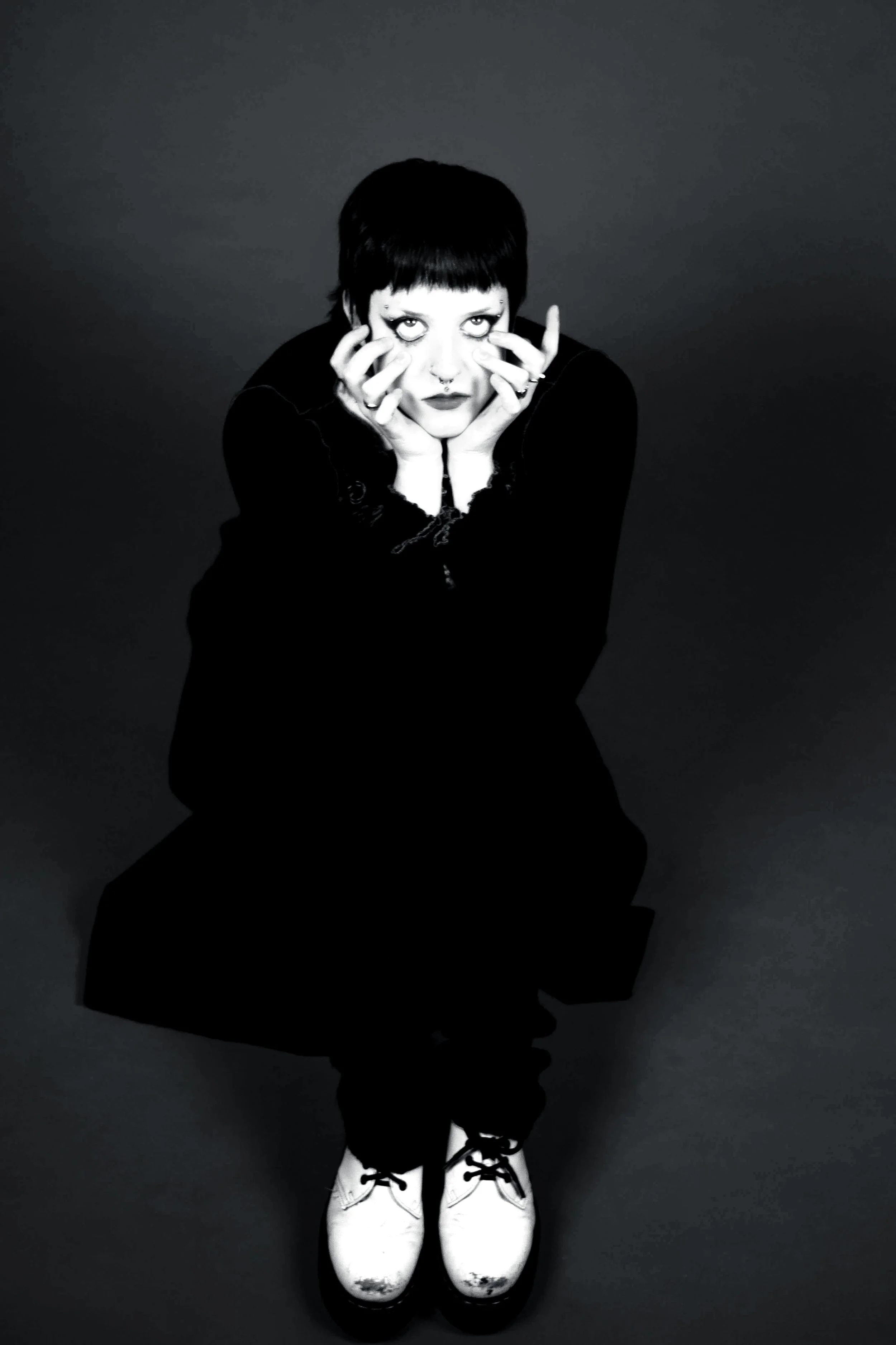 Black and white photo of a person with short dark hair and makeup, sitting on the floor with their head resting in their hands, wearing white boots on a dark background.