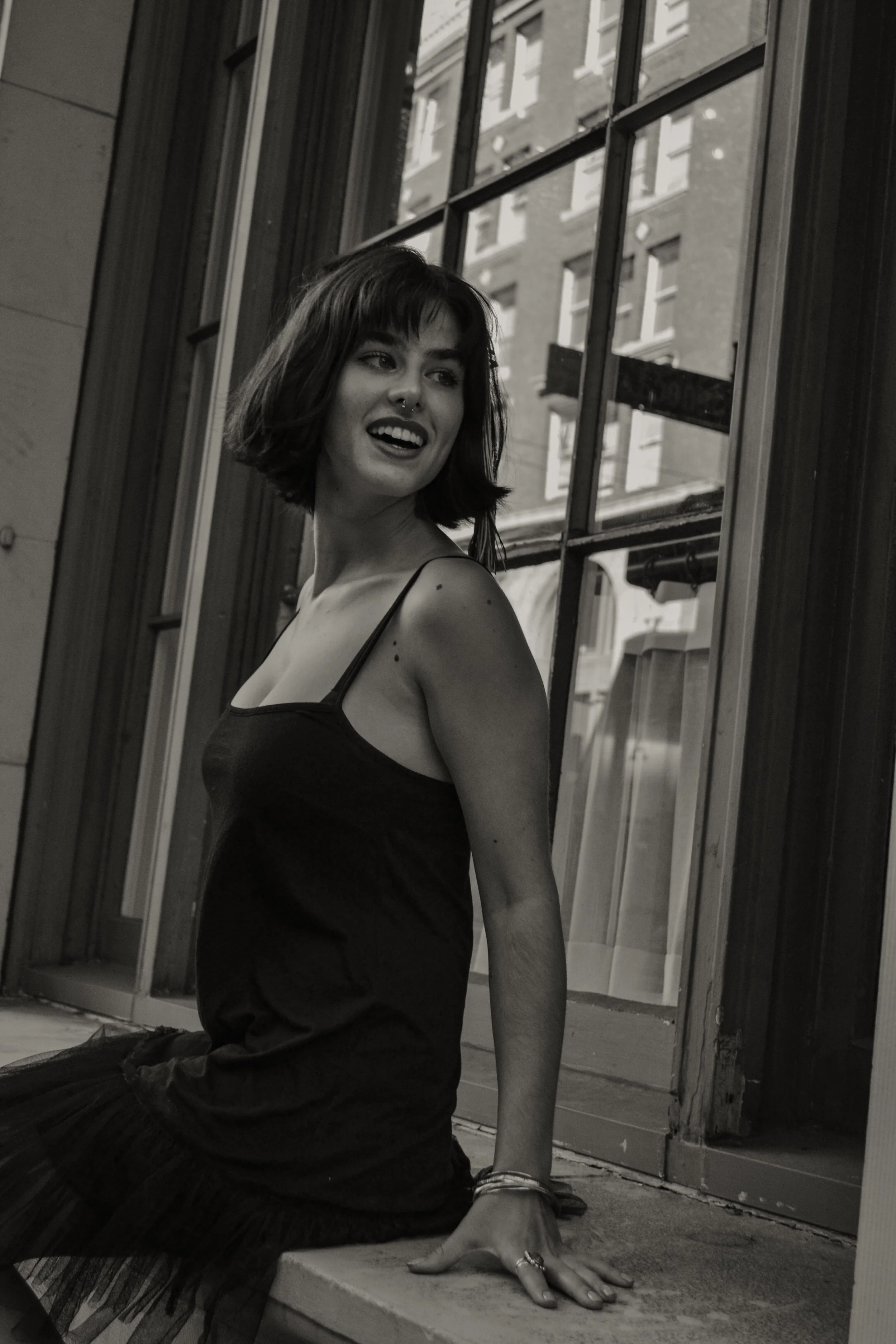 A woman with short dark hair, wearing a black spaghetti strap dress, is sitting by a large window in an urban setting. She has a septum piercing, rings on her fingers, and is looking over her shoulder with a relaxed expression. The background shows a