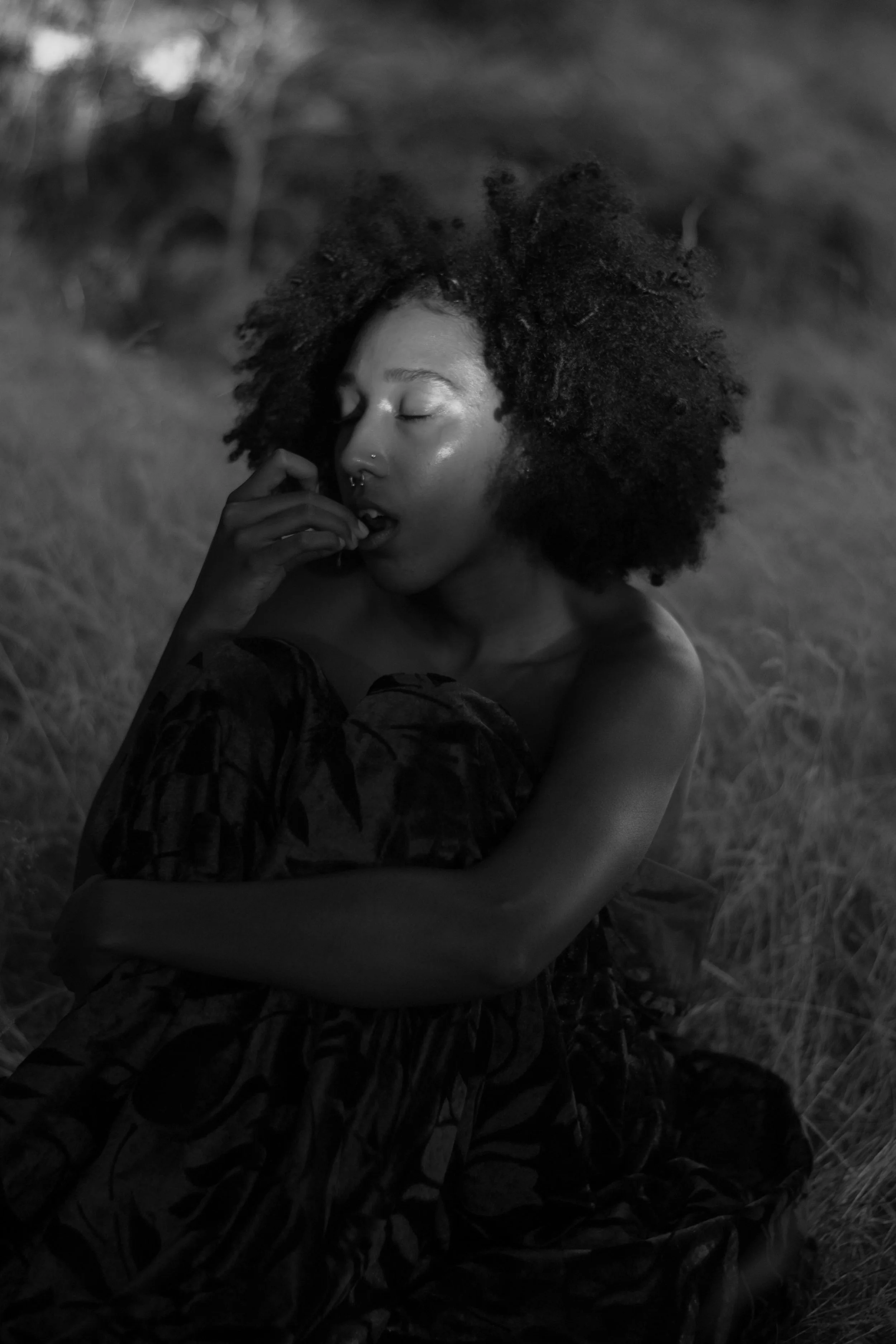 A black and white photograph of a woman with natural curly hair, eyes closed, in a peaceful pose outdoors, with tall grass and blurred trees in the background.