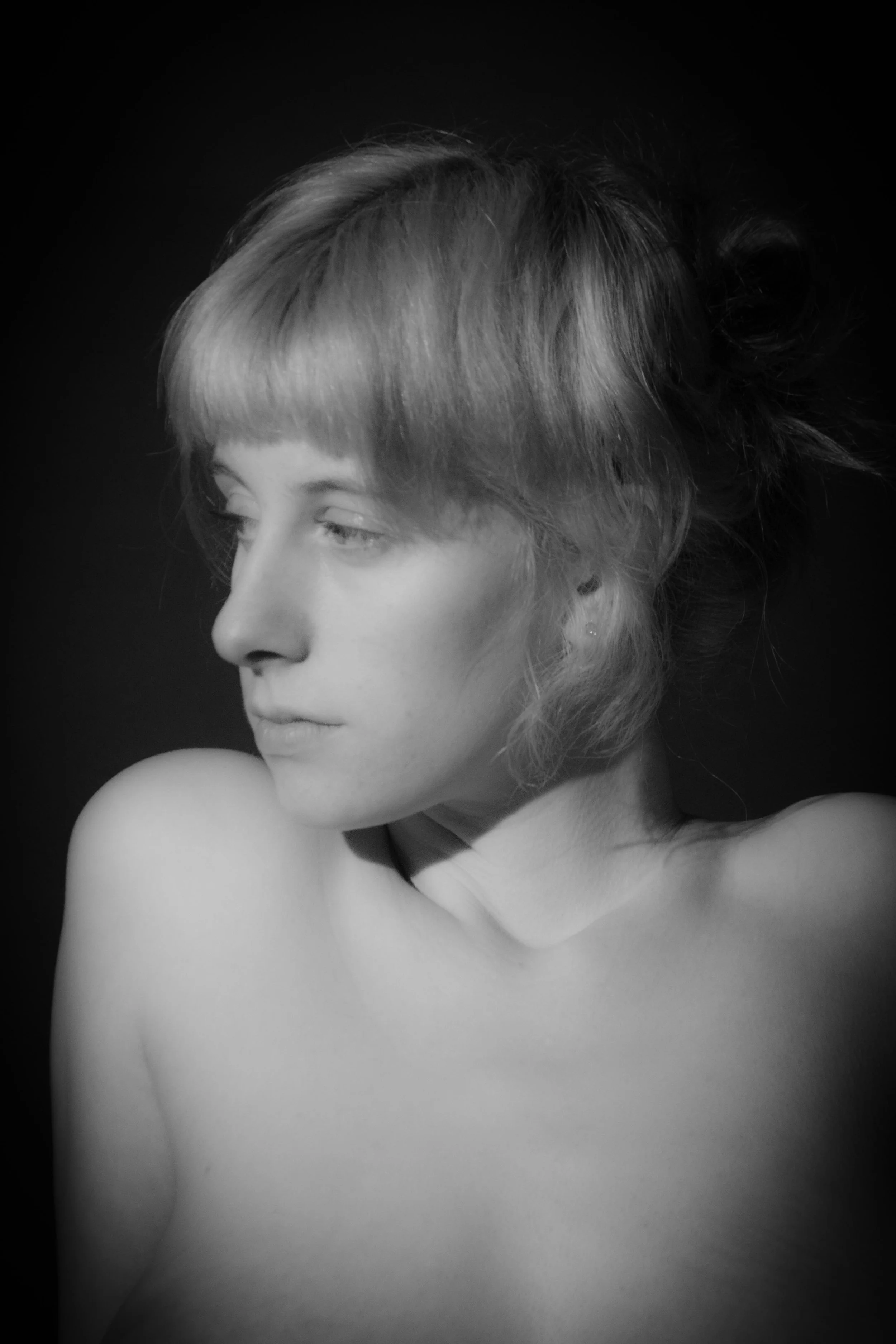 Black and white portrait of a woman with short hair looking down with her shoulder bare against a dark background.