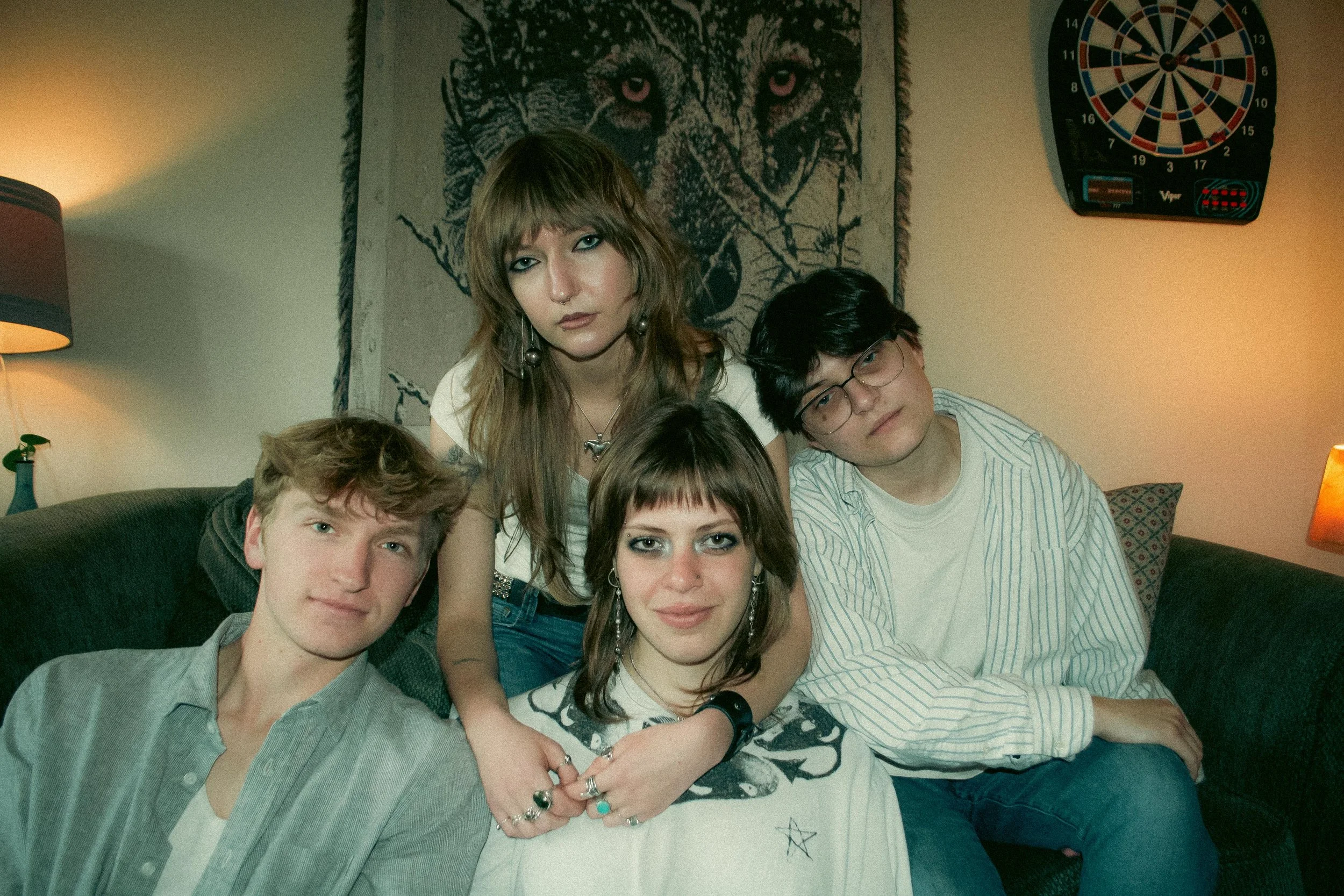 Group of young people sitting on a couch in a cozy room, with a woman with long hair and makeup sitting in the middle, surrounded by friends, with a dartboard and a lion painting on the wall in the background.