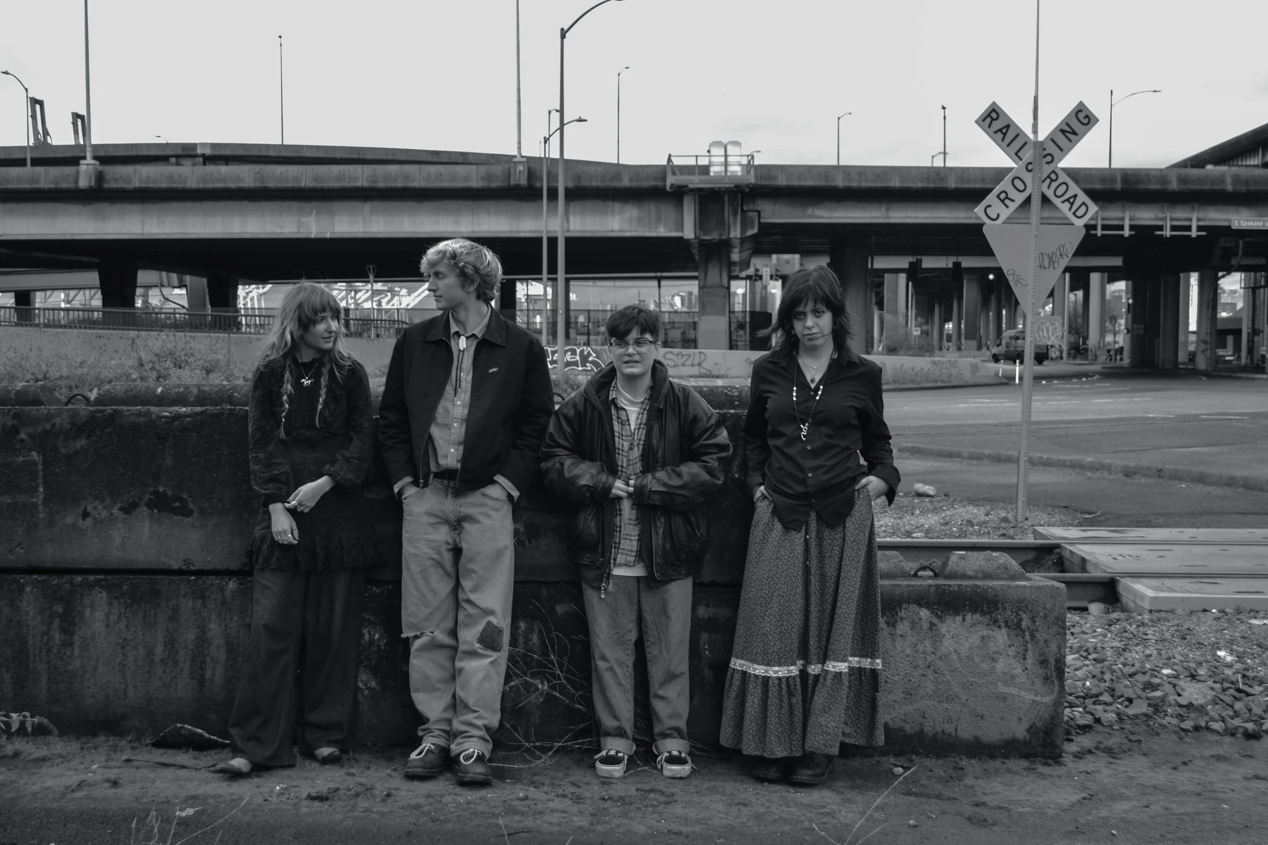 Four young people standing in front of an urban overpass on a city street, with a railroad crossing sign and graffiti visible on the background.