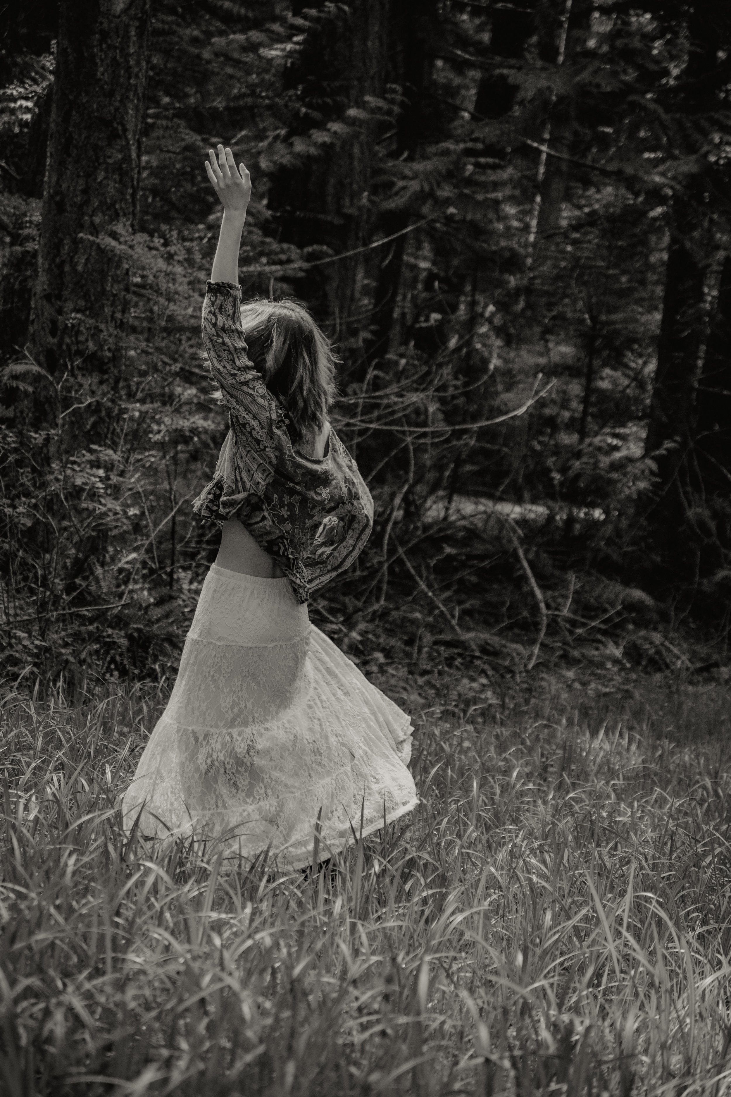 A woman standing in a grassy field with a forest in the background, wearing a lace skirt and a patterned jacket, with one arm raised overhead, in black and white.