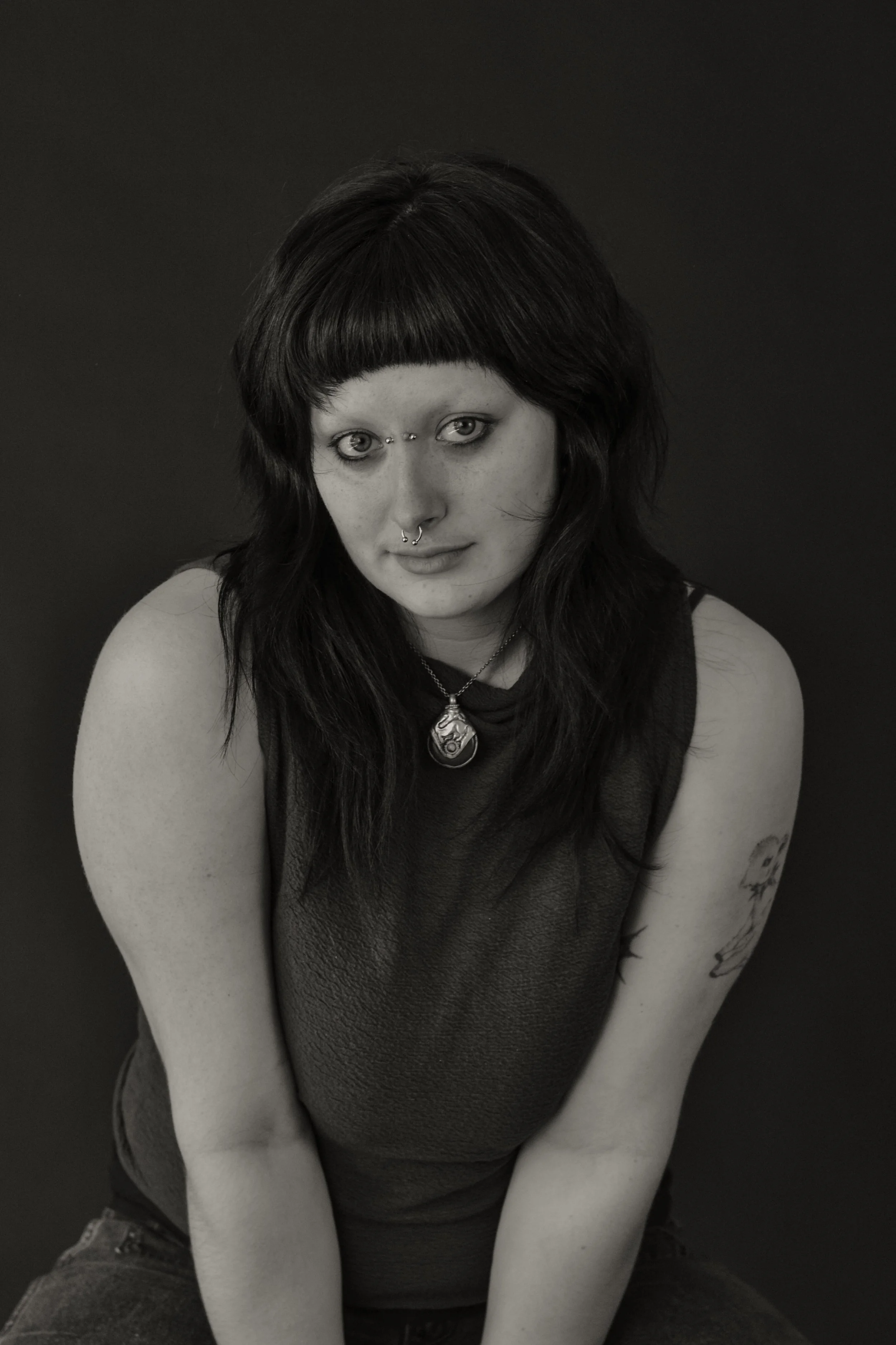 Black and white photo of a young woman with dark hair and bangs, wearing a ring on her nose, a pendant necklace, a sleeveless top, and tattoos on her arms, sitting with her arms crossed in front of her.