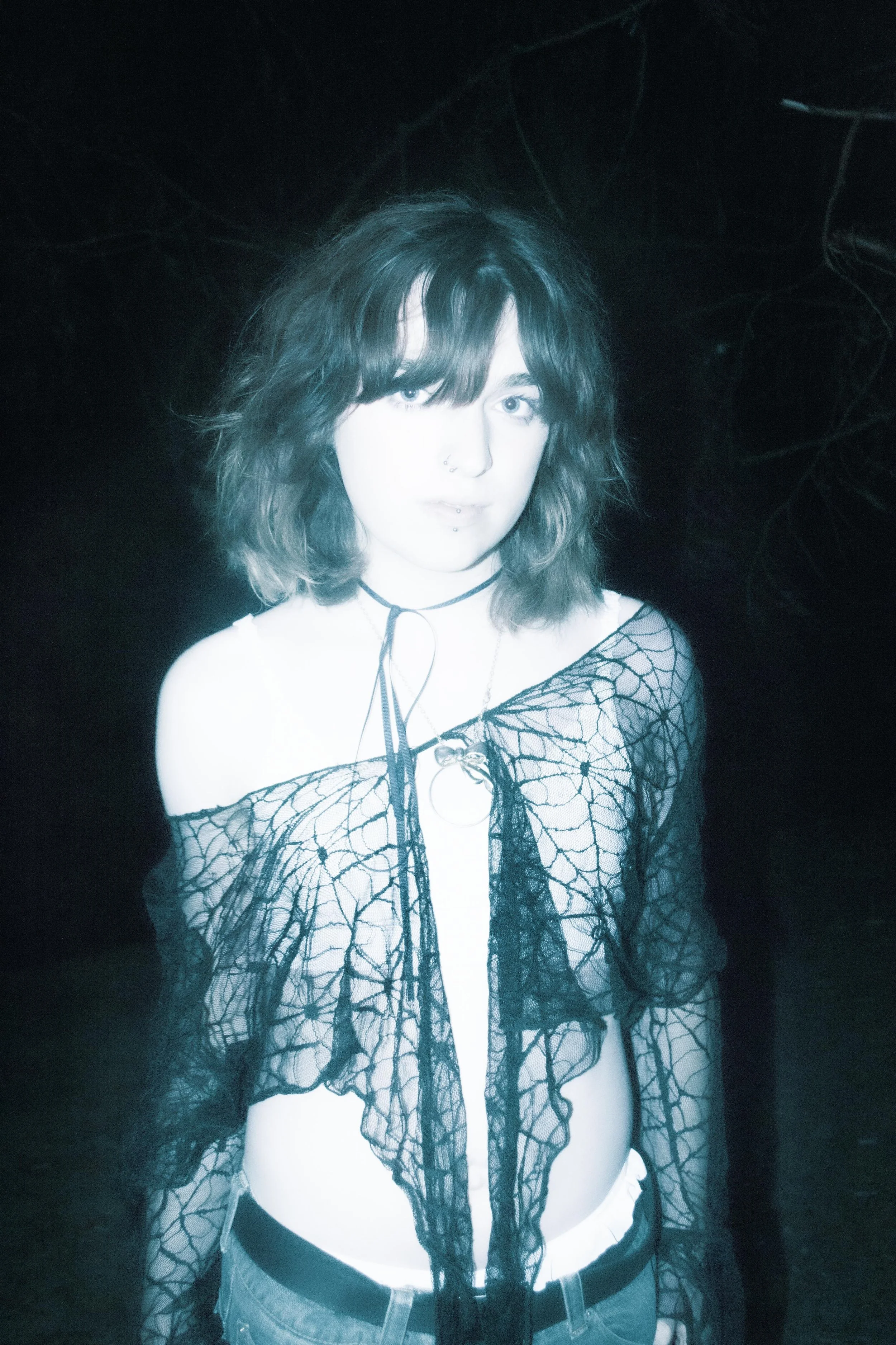 Person with wavy shoulder-length hair wearing a black lace top, choker necklace, and white pants, standing against a dark background with faint web-like patterns.